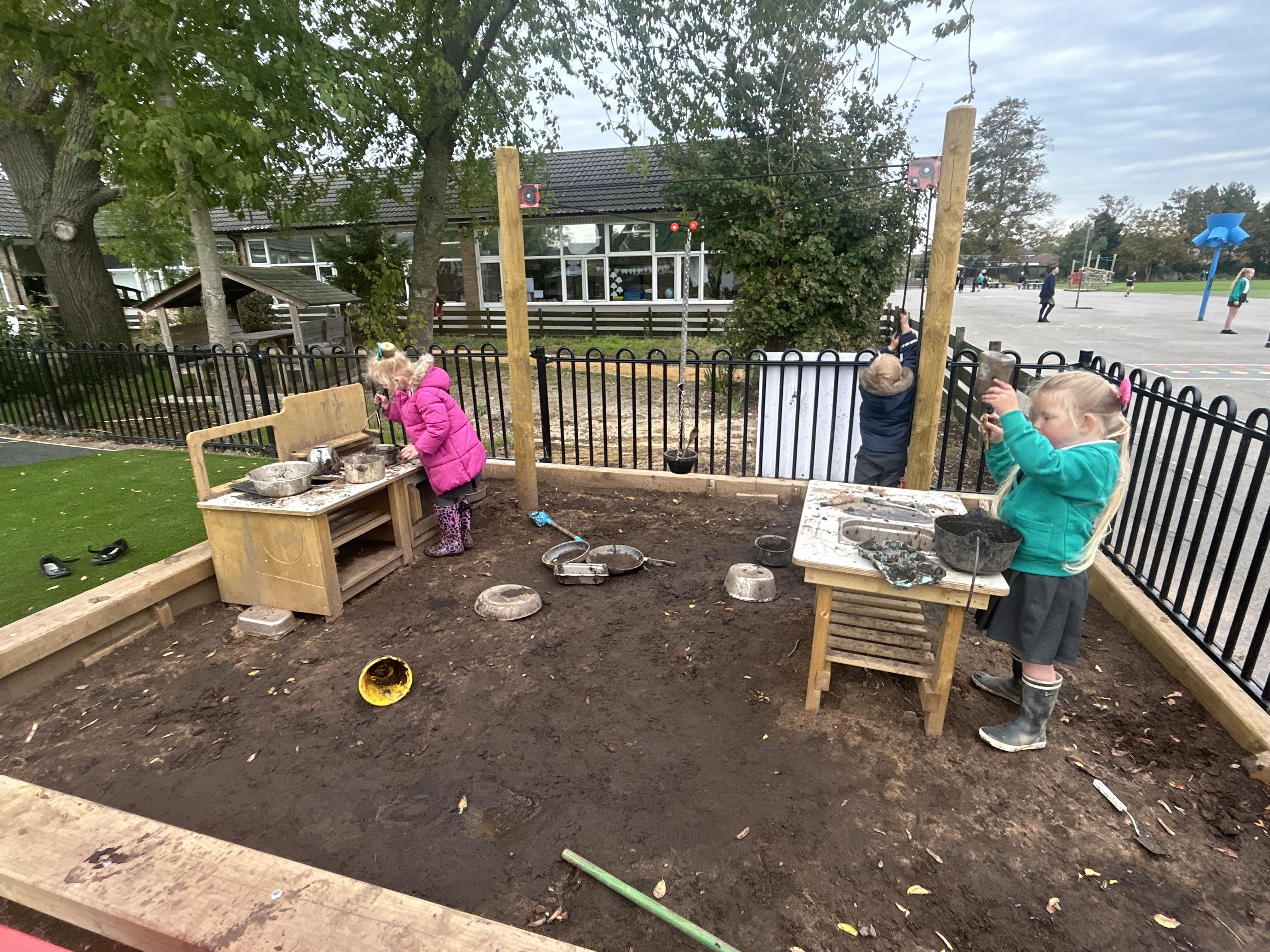 Three children are playing with Messy Play equipment in a muddy area. All three children are wearing coats and wellies, as puddles form in the mud