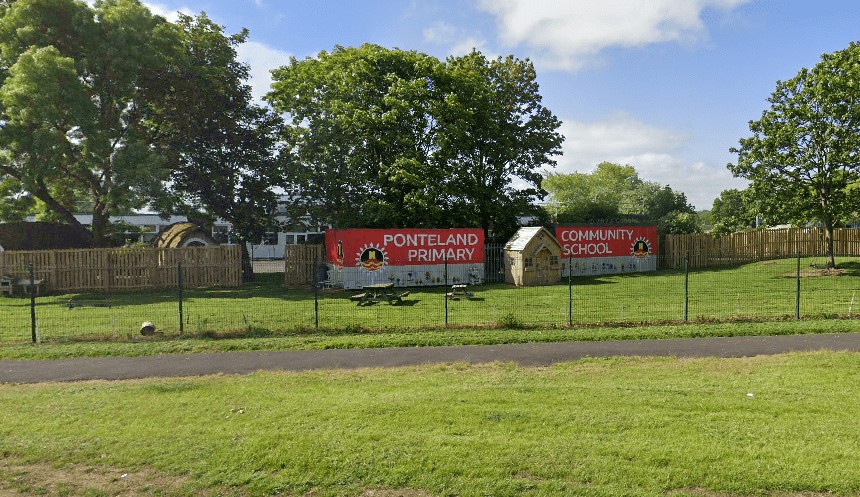 A field of grass with two big posters up saying "Ponteland Community Primary School". A mesh fence can be seen going around the area and securing the school.