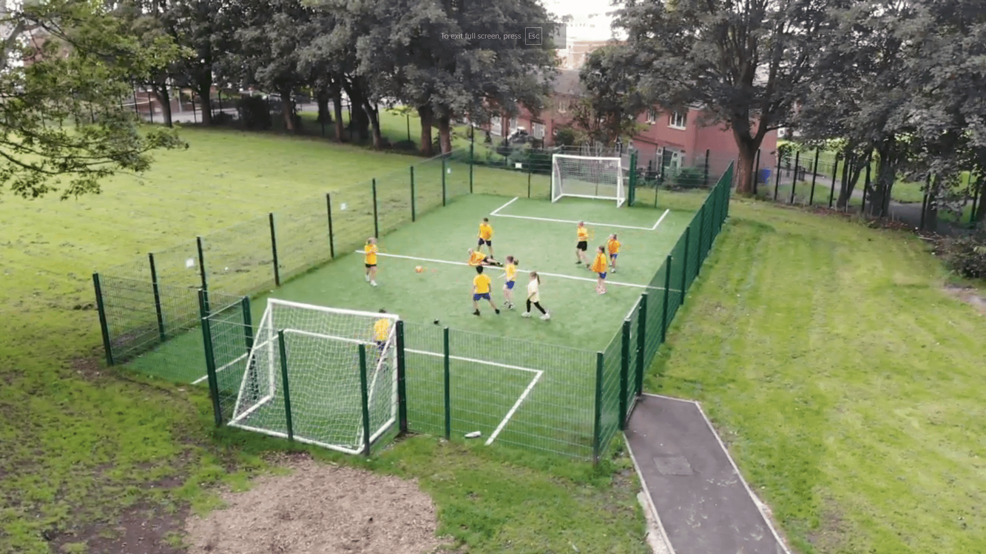 A high angle photo that showcases a football match being played on an Artificial Grass MUGA. A teacher can be seen being a referee as the children are in their own teams.