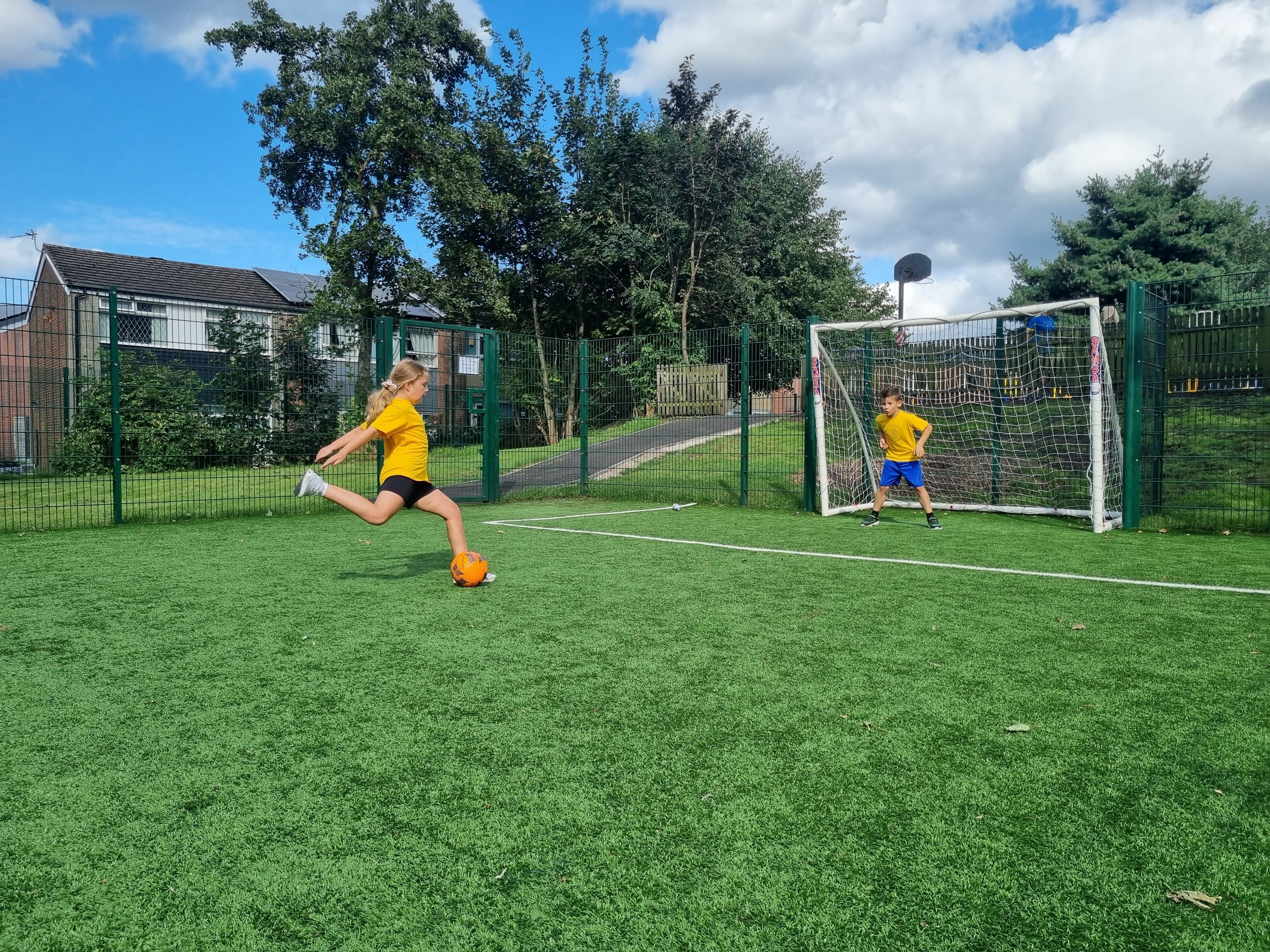 A child is taking a penalty kick against another child who is in the recessed goal that has been created within the MUGA fencing.