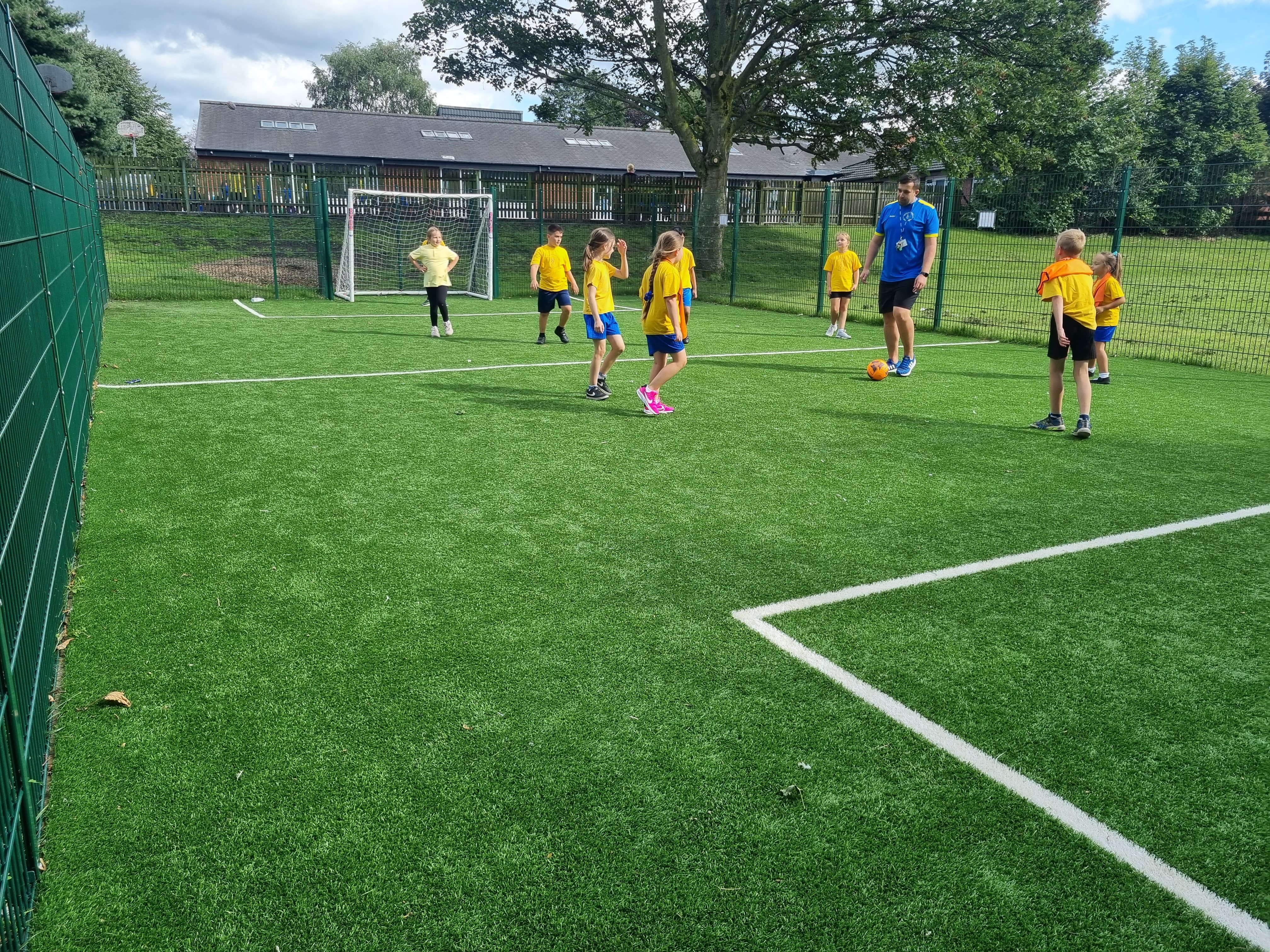 A group of children are playing football on a MUGA. Half the children are wearing orange bibs and a teacher can be seen wearing a blue top and black shorts.