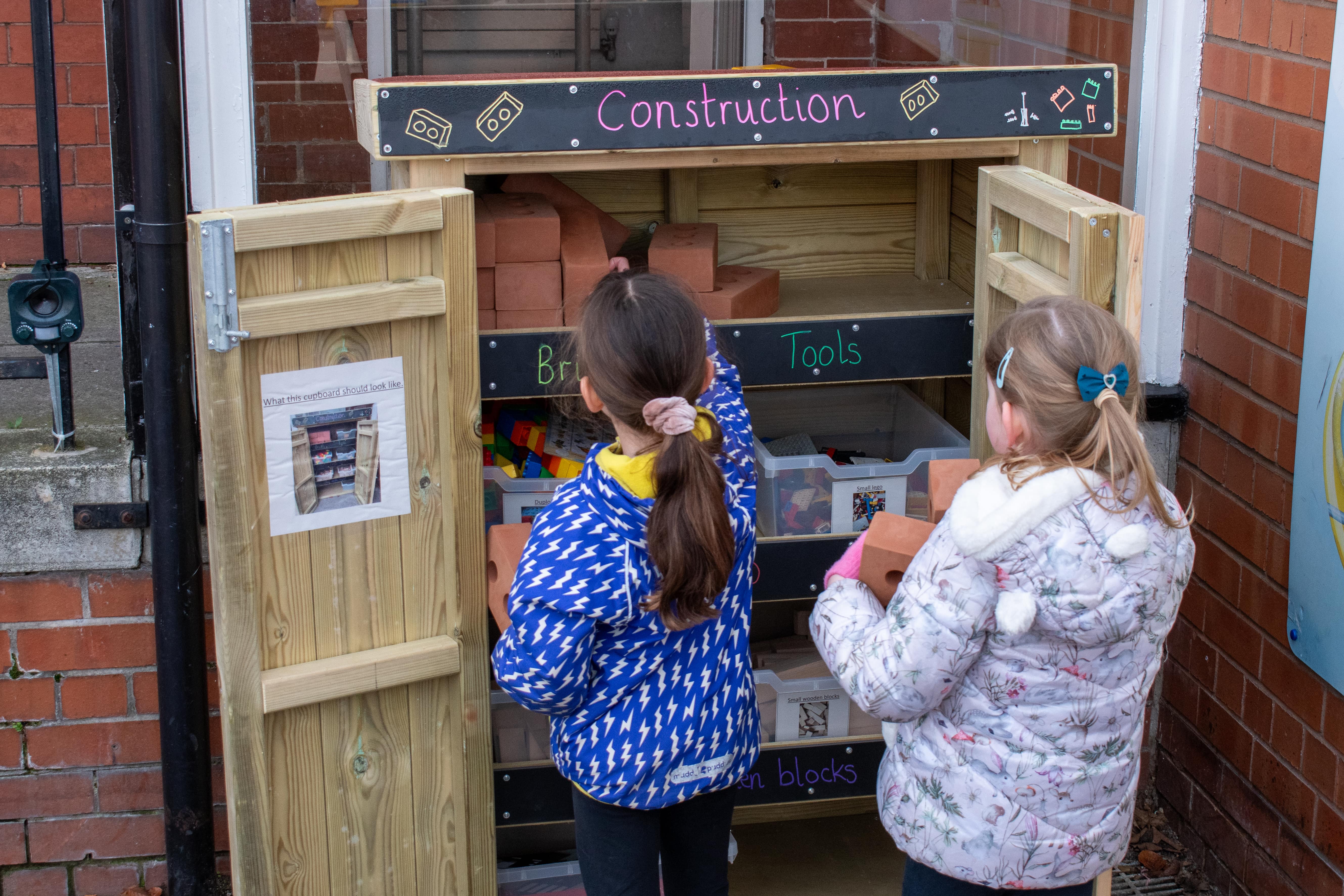 Two children are looking through a storage unit with the title "Construction", which contains a variety of construction play resources. The children are retrieving different items from the unit.  