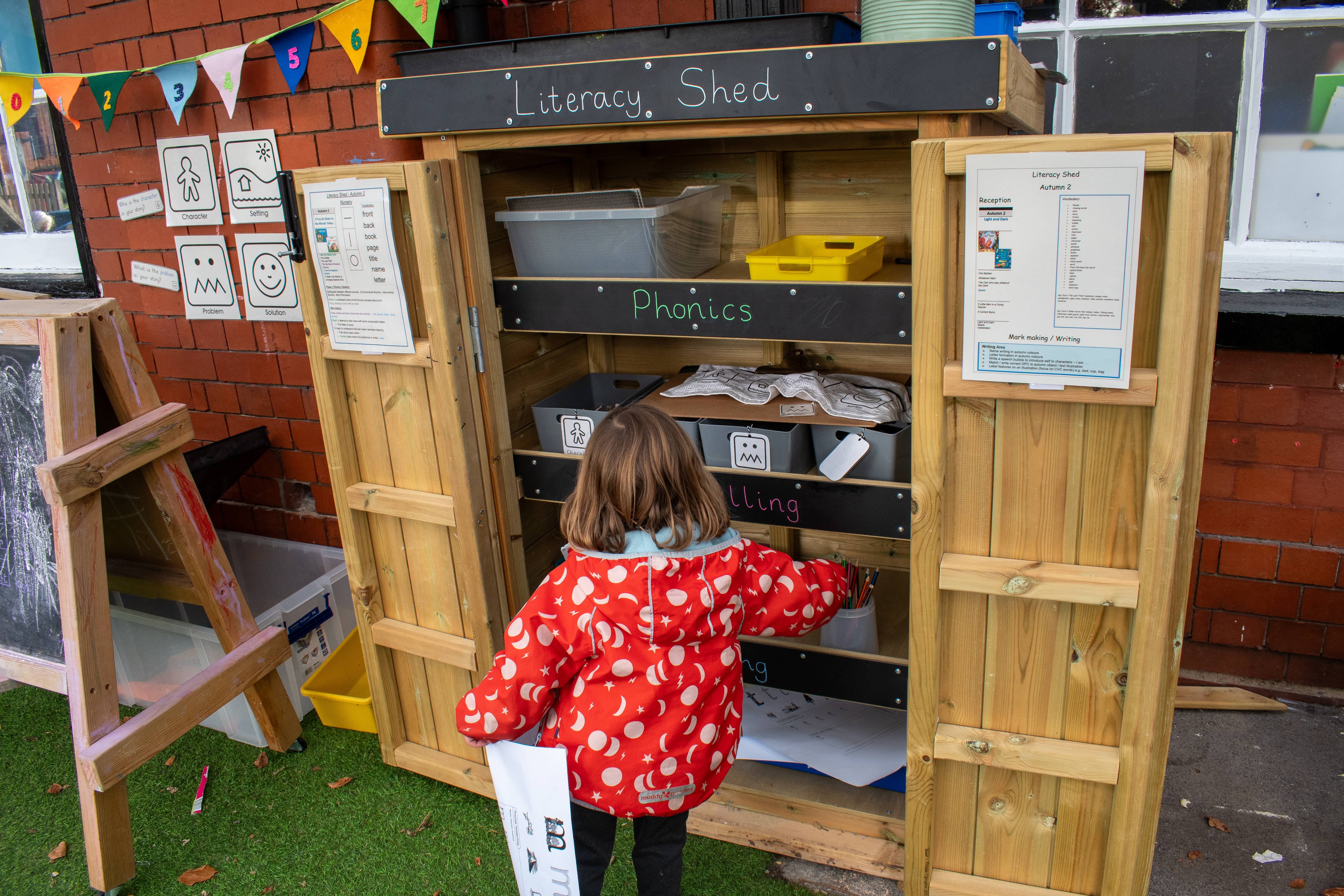 A little girl wearing a spotty coat is looking through an Acorn Self-Select Store that has the title "Literacy Shed". The little girl is grabbing some paper and other resources from the shed.