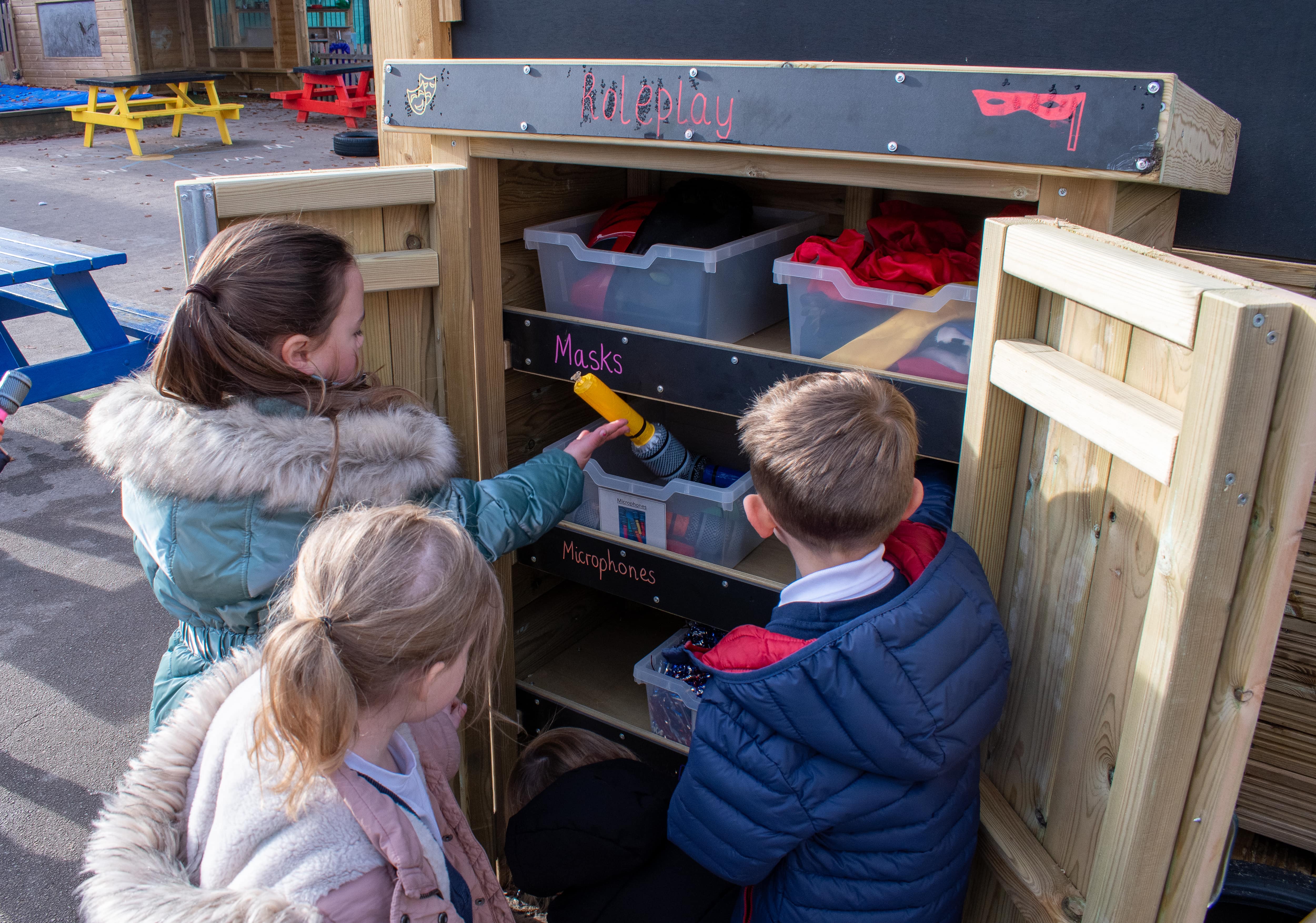 Three children are looking through an Acorn Self-Select Store unit which has the title of Roleplay. The children are holding role play items.