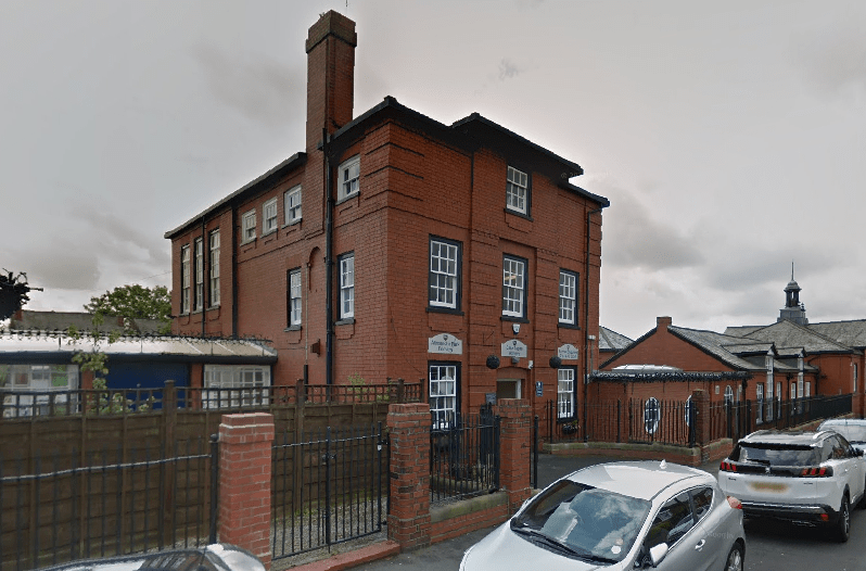 A red brick building that has signs attached to the building saying "Alexandra Park Primary School". A bunch of cars are parked alongside the road.