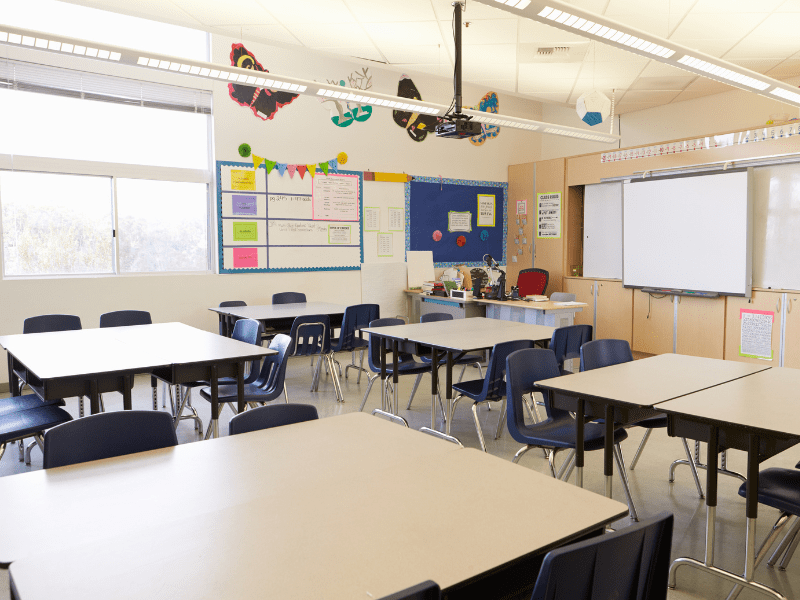 An empty classroom which has a variety of tables and chairs scattered across the room. A display board and whiteboard can be seen in the far background.