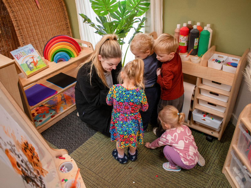 A nursery practitioner is kneeling on the floor to talk to four EYFS children. The children look engrossed in their project.