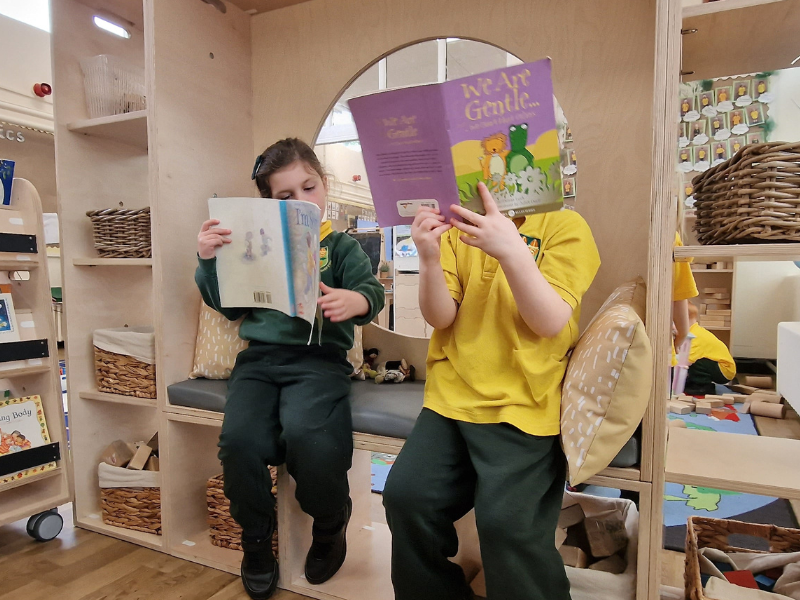 Two children are sat in a classroom and are both reading different books.