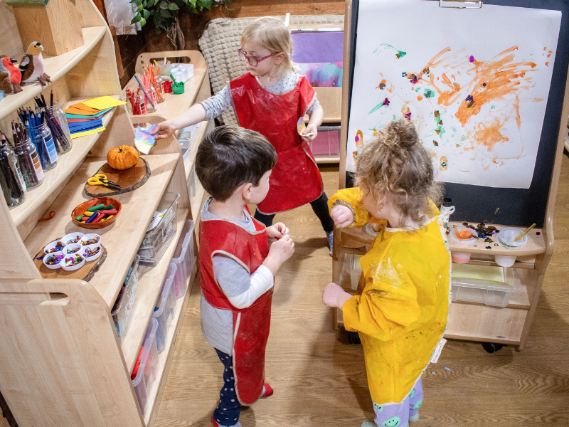 Three children are stood around in a creative area and are using a variety of arts and crafts materials. The children are drawing on the art easel and having fun.