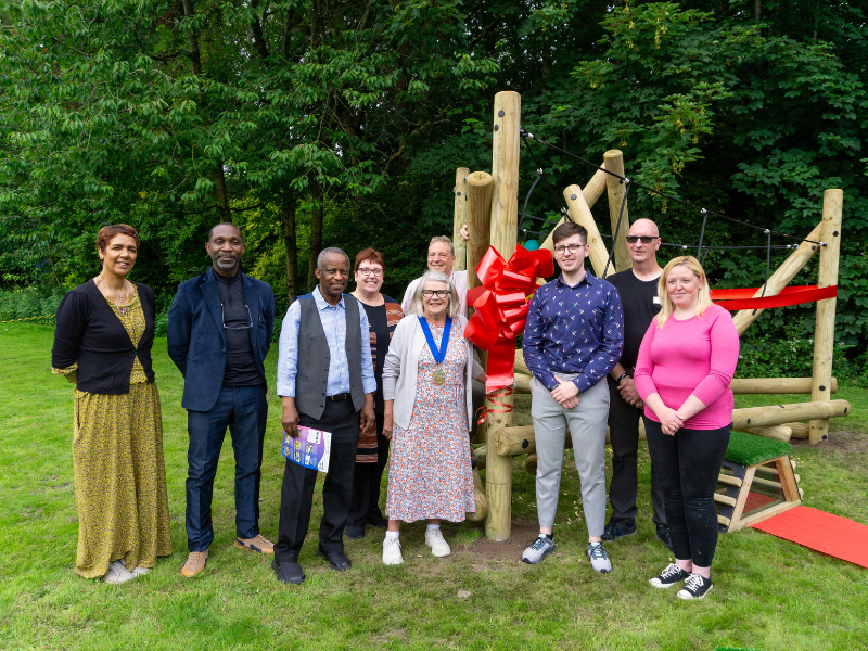 A group of adults are stood beside a piece of play equipment that has a red ribbon wrapped around it. Some members of the party are from the local authority.