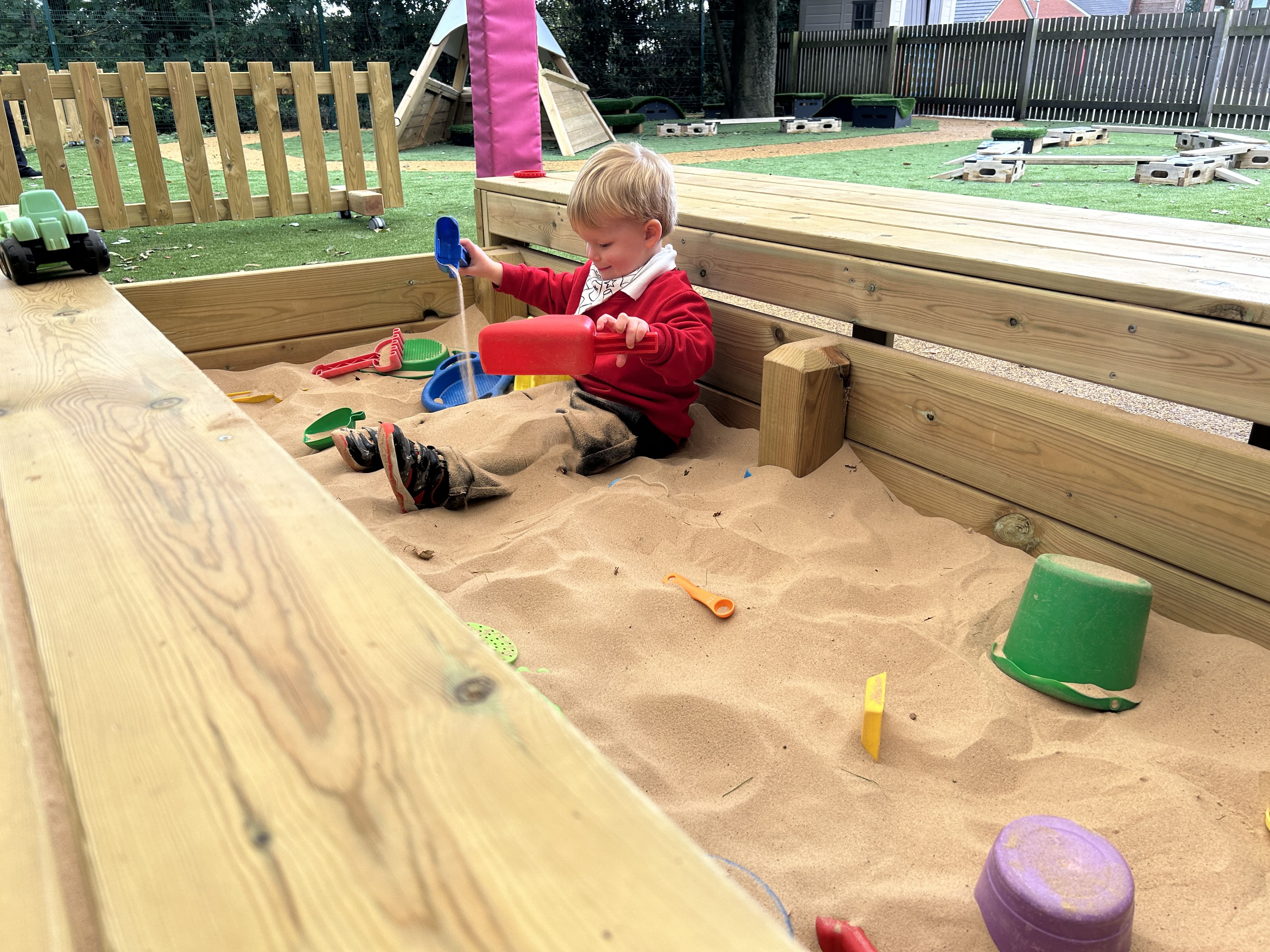 A little boy is sat in a sandpit as he digs up sand on his shovel and pours it on his legs. He looks happy as he engages in sensory play.