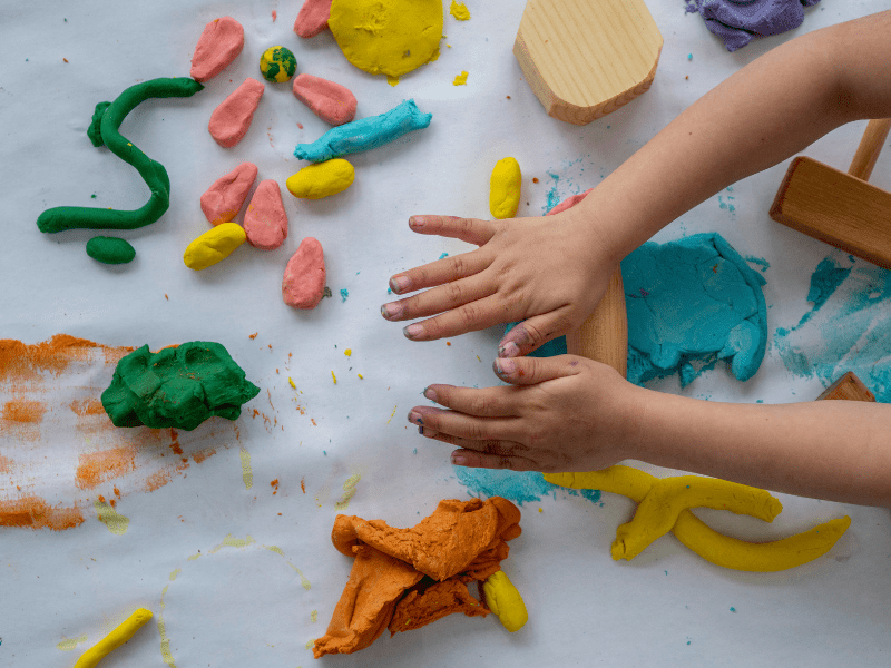 A child's hands can be seen on a table as they hold some playdough. They are moulding the dough into different shapes and items.