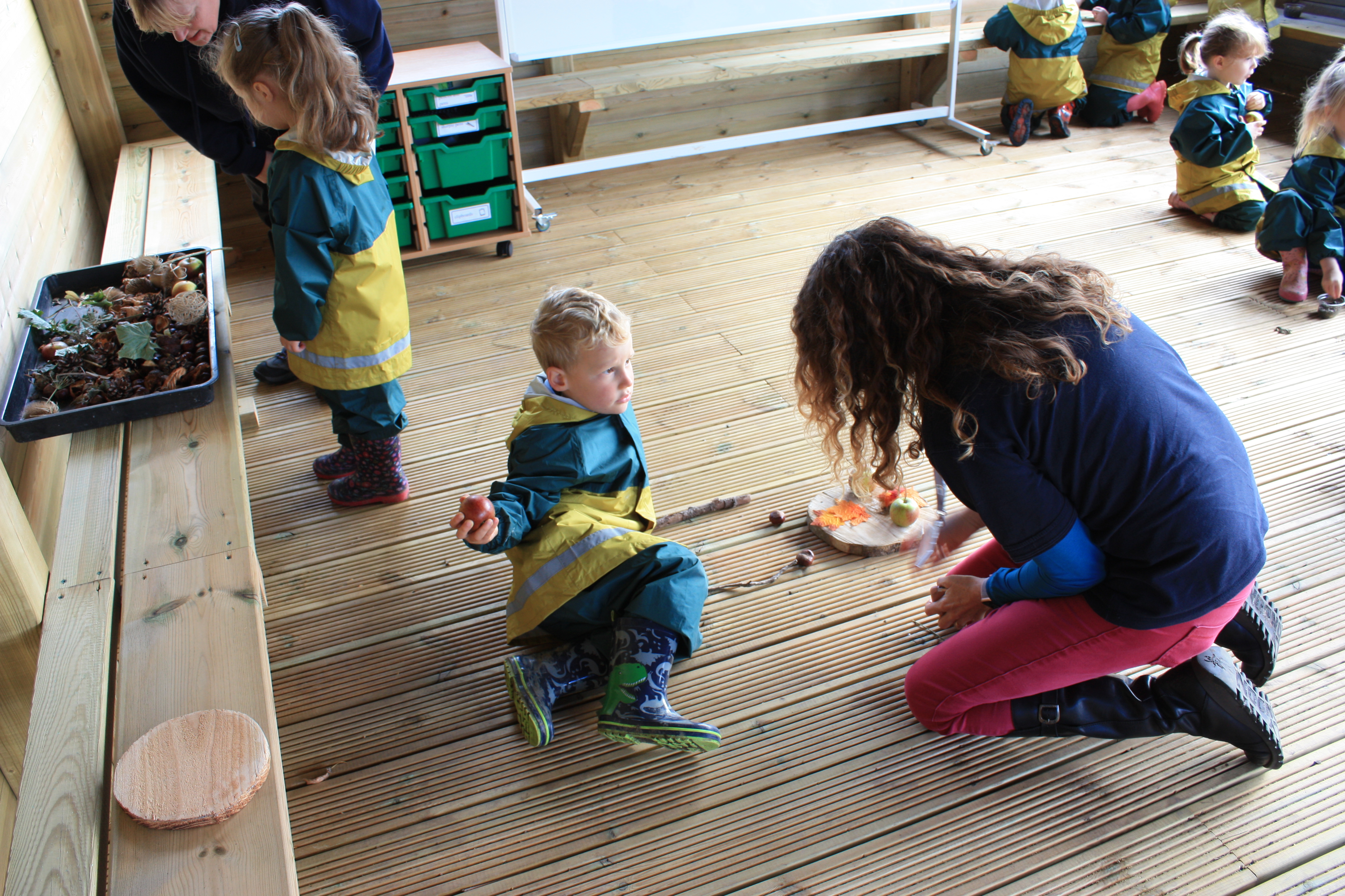 A little boy is lying on an outdoor classroom floor as a teacher kneels next to him, presenting him with a variety of natural materials to promote sensory play.