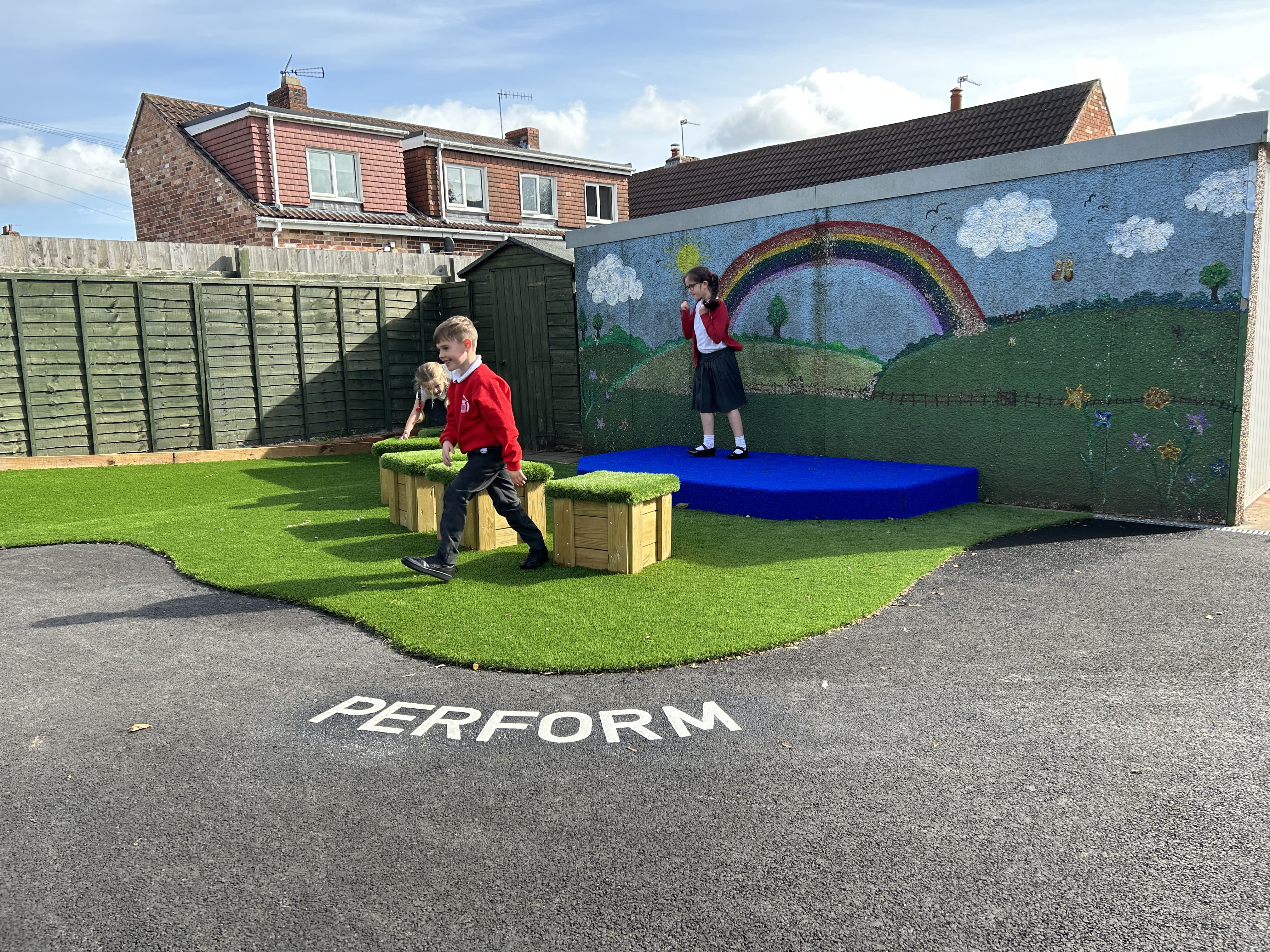 A little girl is stood on a blue performance stage as two pupils are moving the artificial grass topped seats.