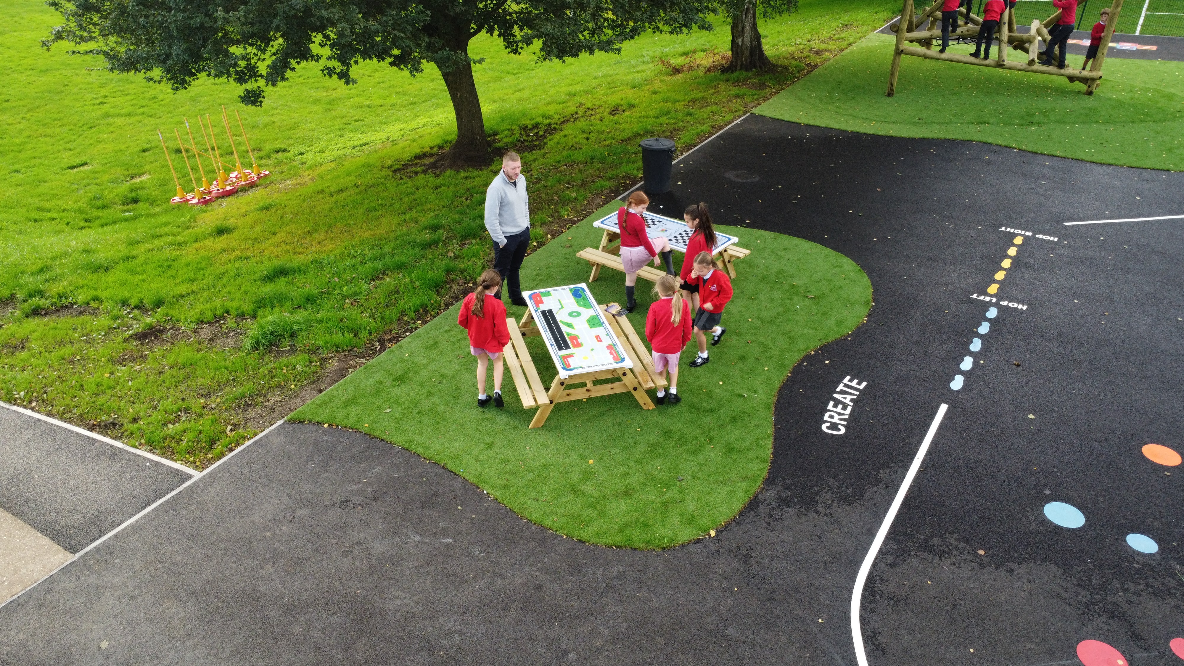 A small group of children are sat on Picnic tables which have different games on the table tops. A teacher is stood beside them.