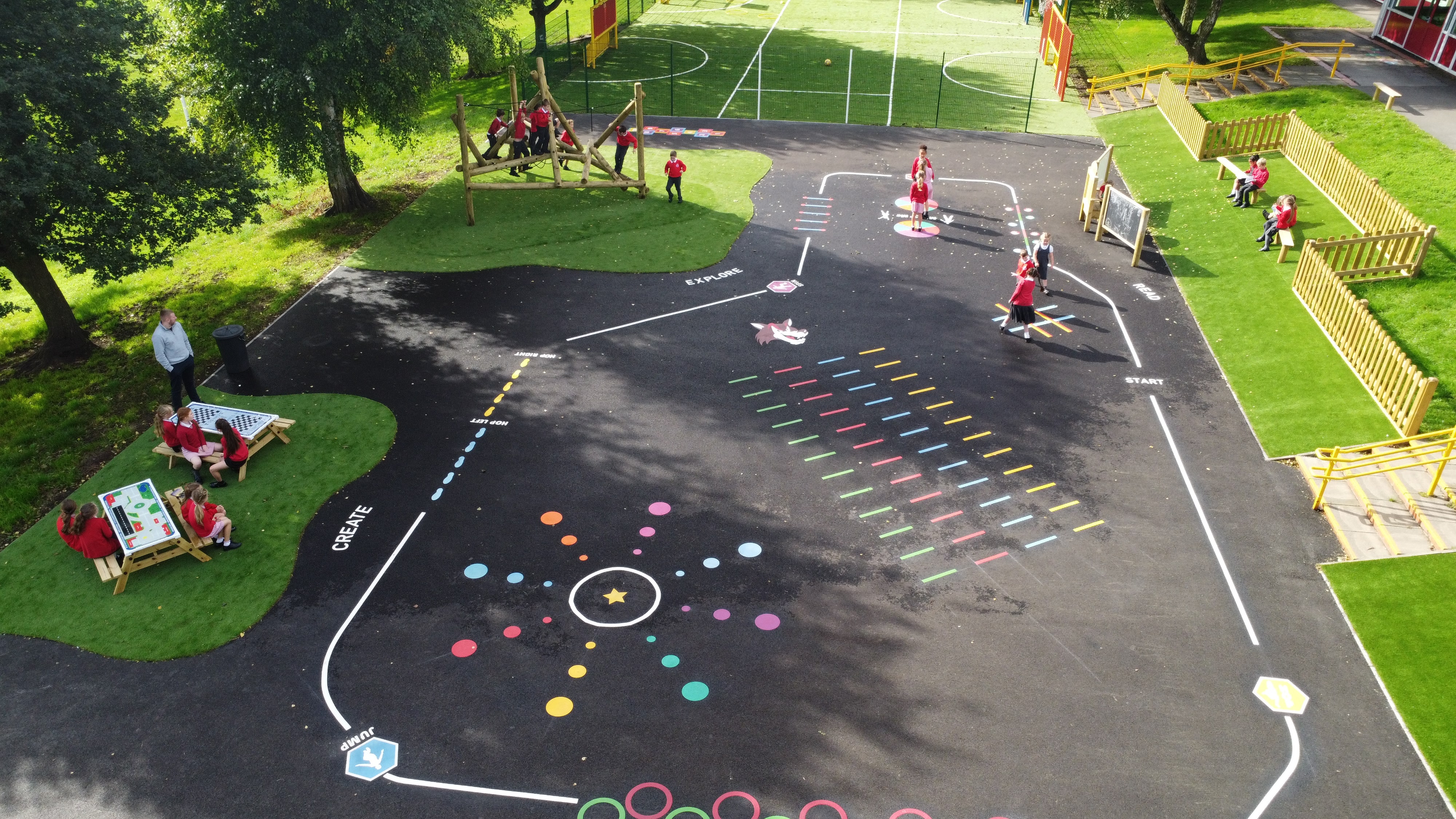 A tarmac playground with a variety of thermoplastic playground markings on it and areas of artificial grass containing different pieces of play equipment.