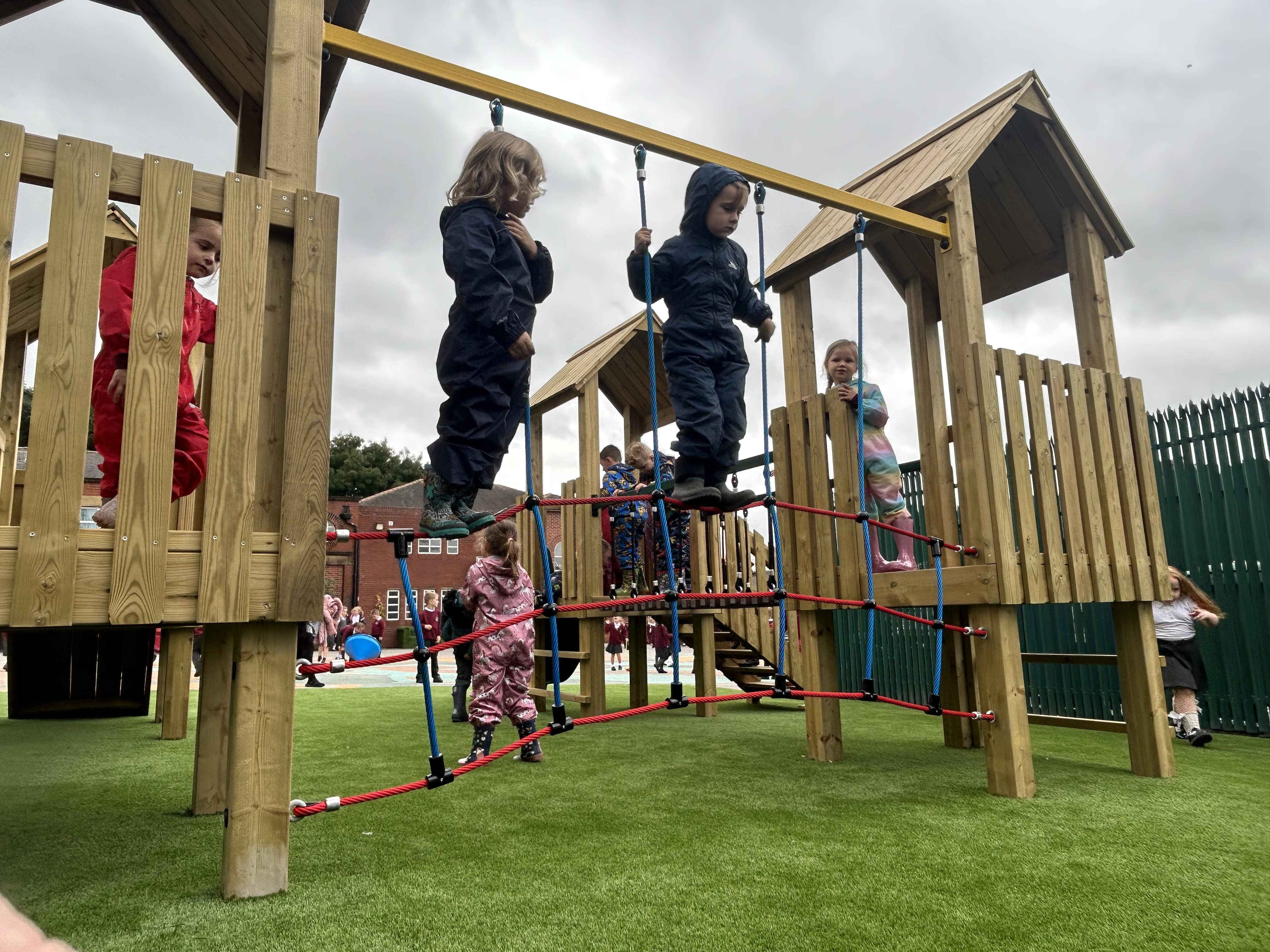 A few children are crossing over a climbing net that is connected between two vertical wooden beams.