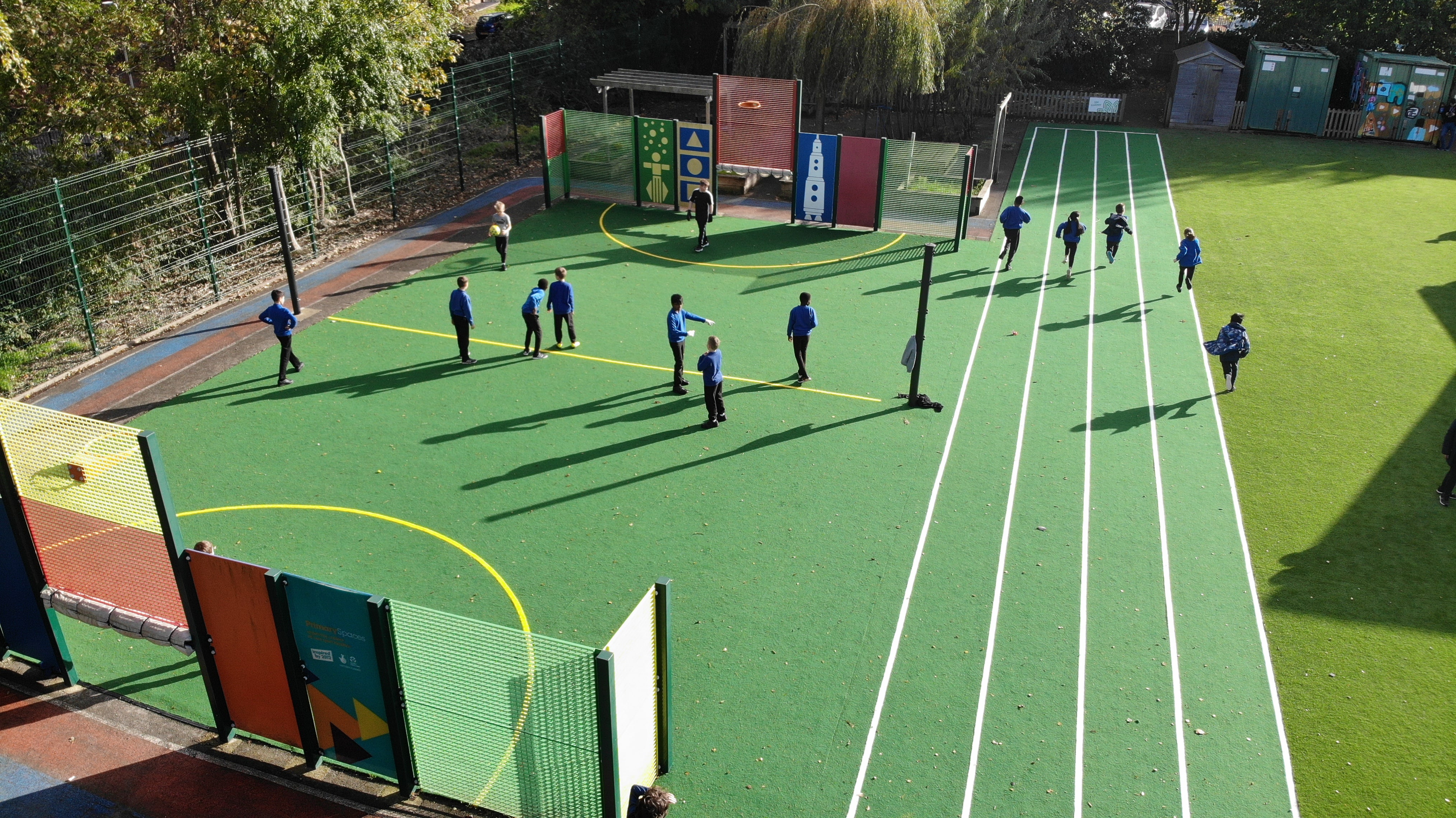 A MUGA court with a running track installed next to it. Both of them are green and have children using them.