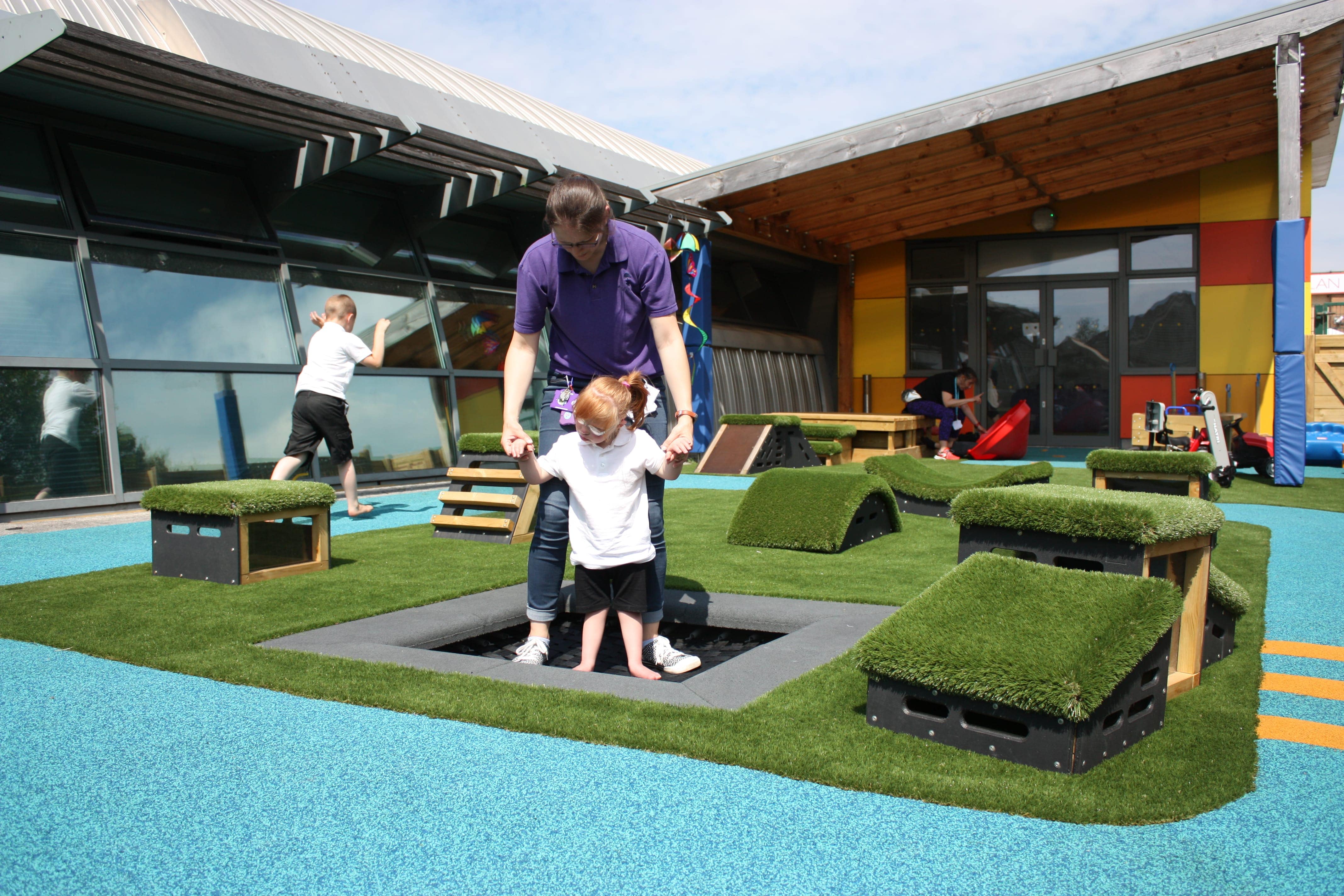 A little SEN boy is bouncing on an in-ground trampoline, with the help of a teacher