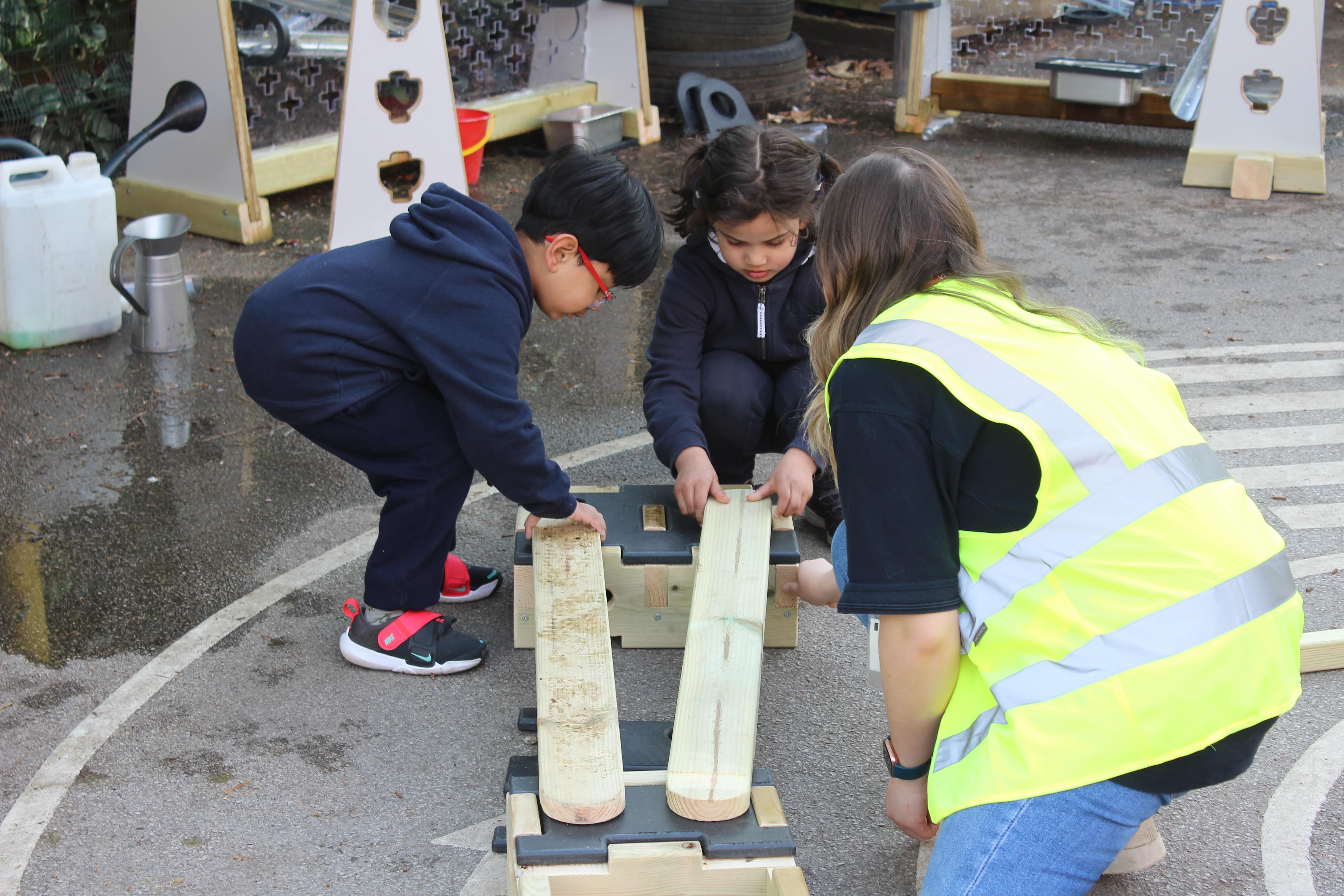 2 SEN Children are placing wooden planks on wooden blocks as an adult in a high-vis jacket is watching and talking to them.