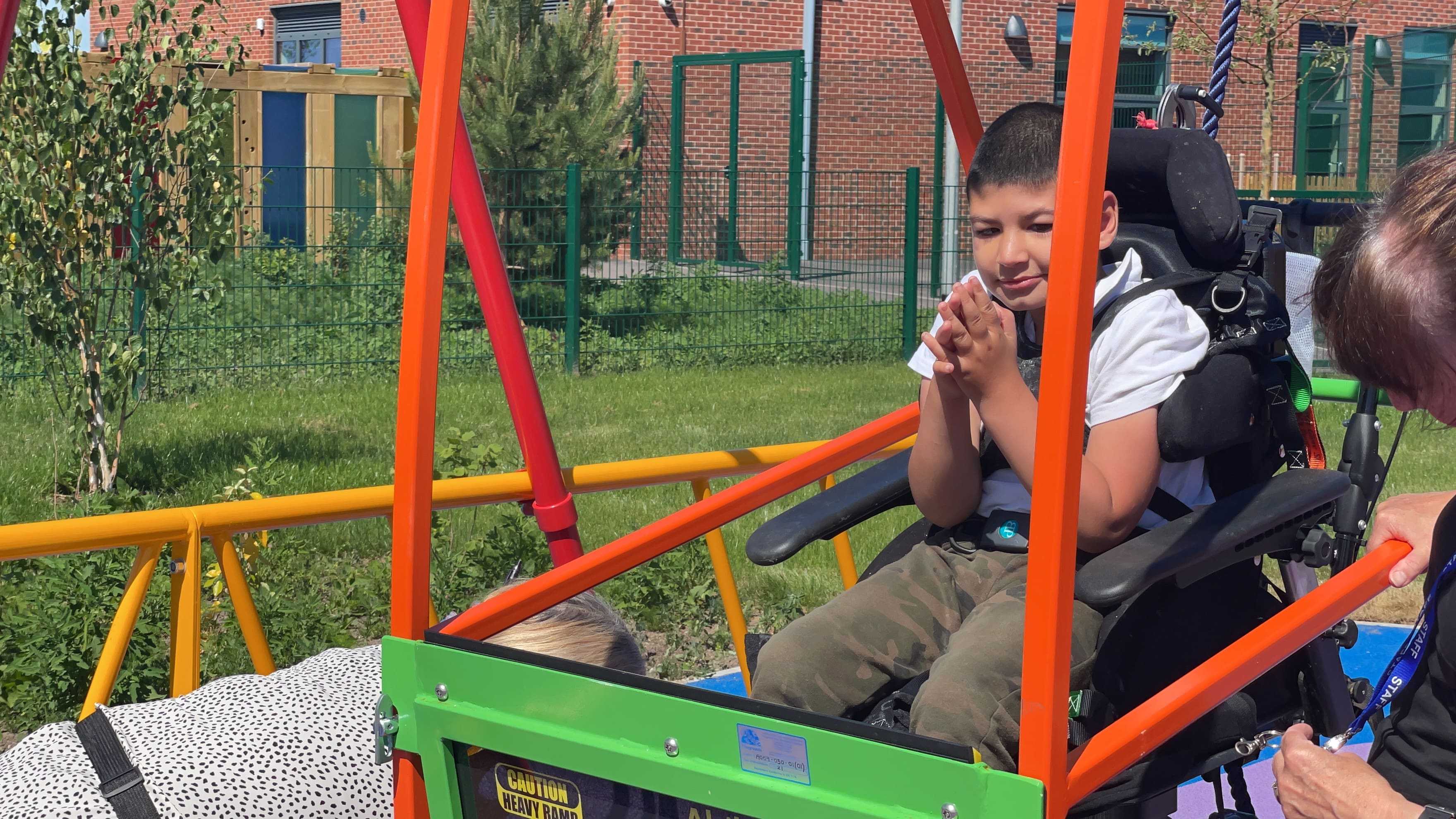 A SEN child is on a brighly coloured wheelchair-accessible swing, with a teacher kneeling next to him.