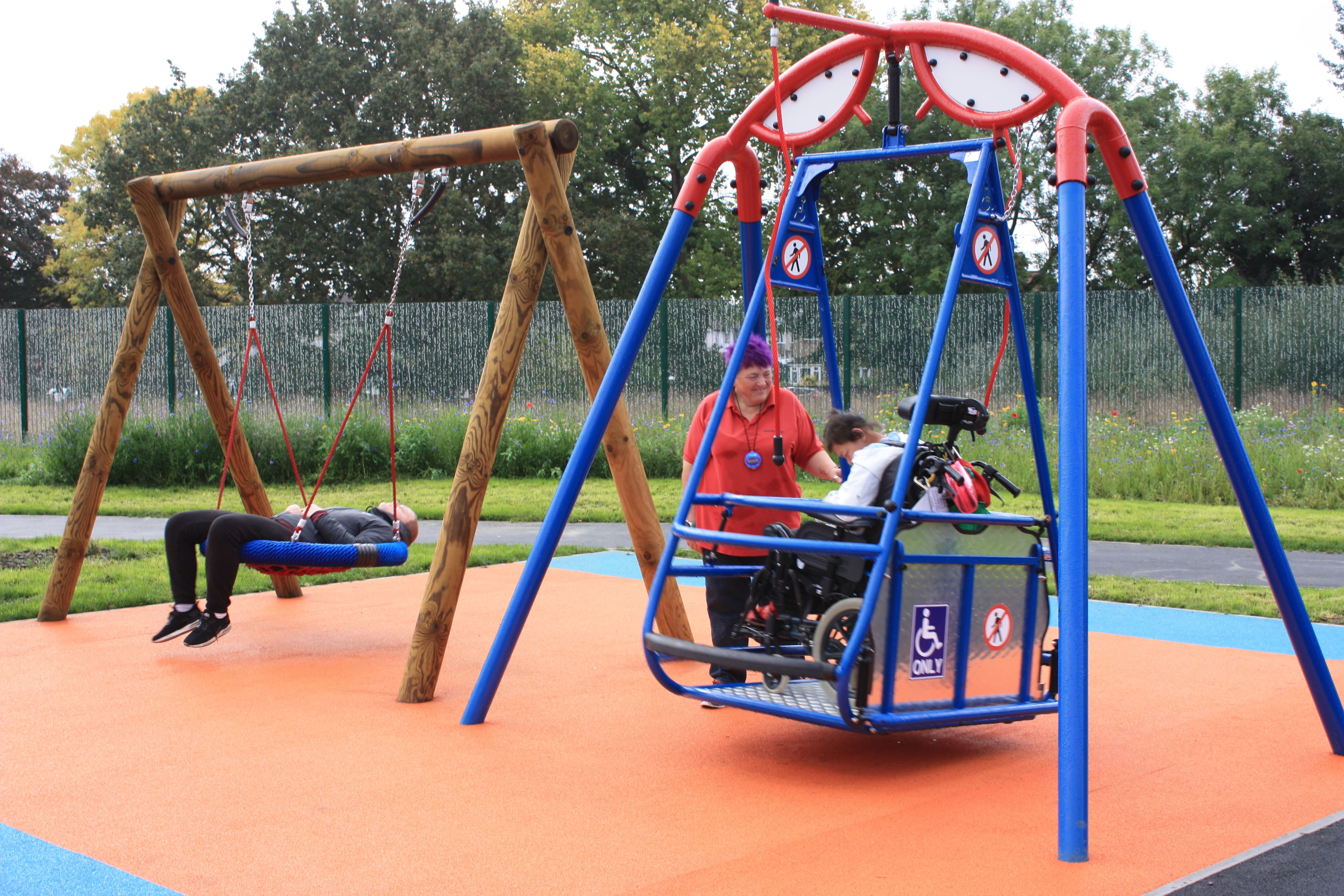 A young SEN boy is on an inclusive wheelchair swing, with his teacher rocking the swing slowly. Another boy can be seen lying down in a basket swing.