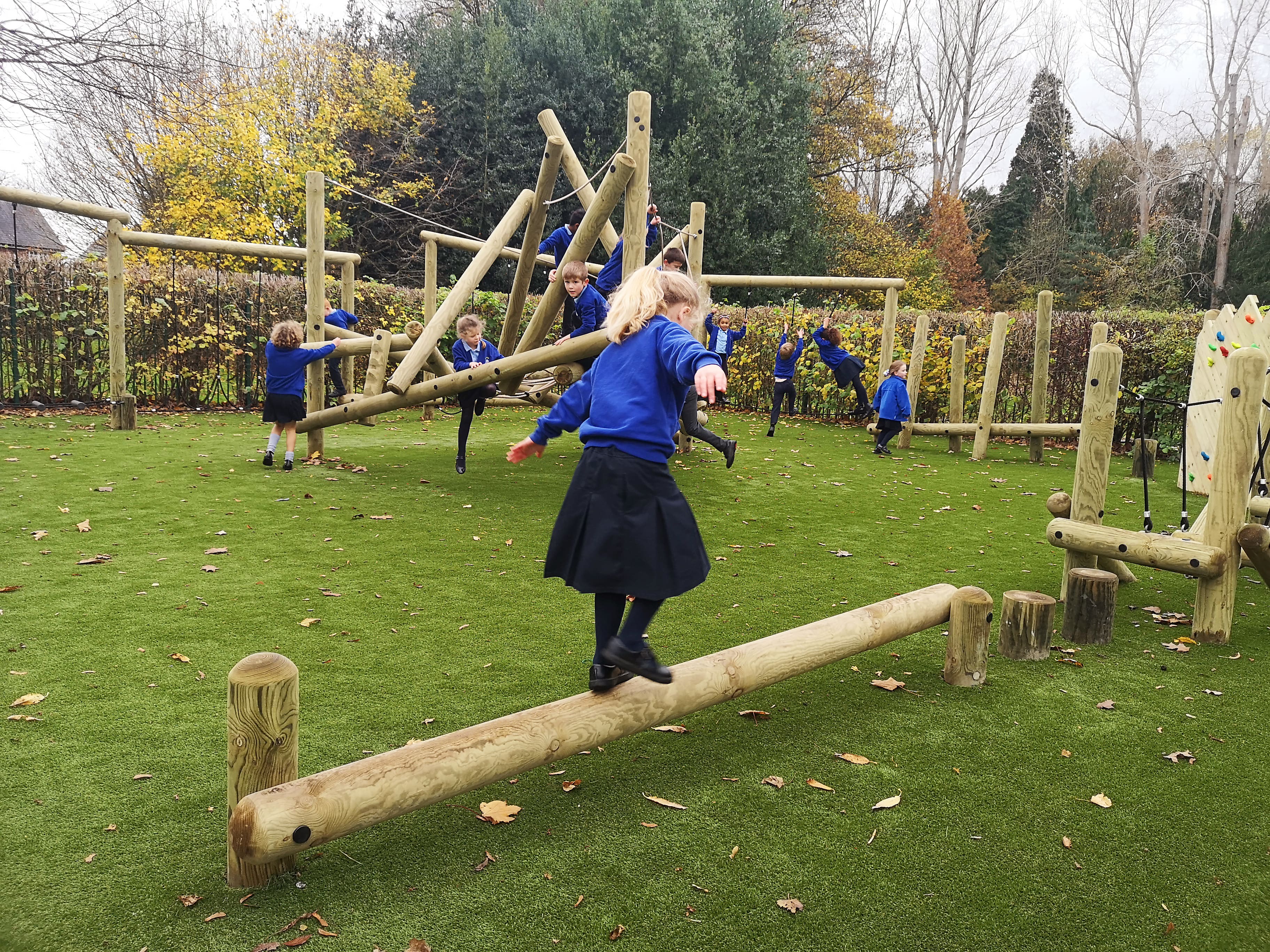 A little girl is traversing over a balance beam, which is a part of a huge trim trail. A group of children can be seen on the other end, climbing on a big climbing frame.