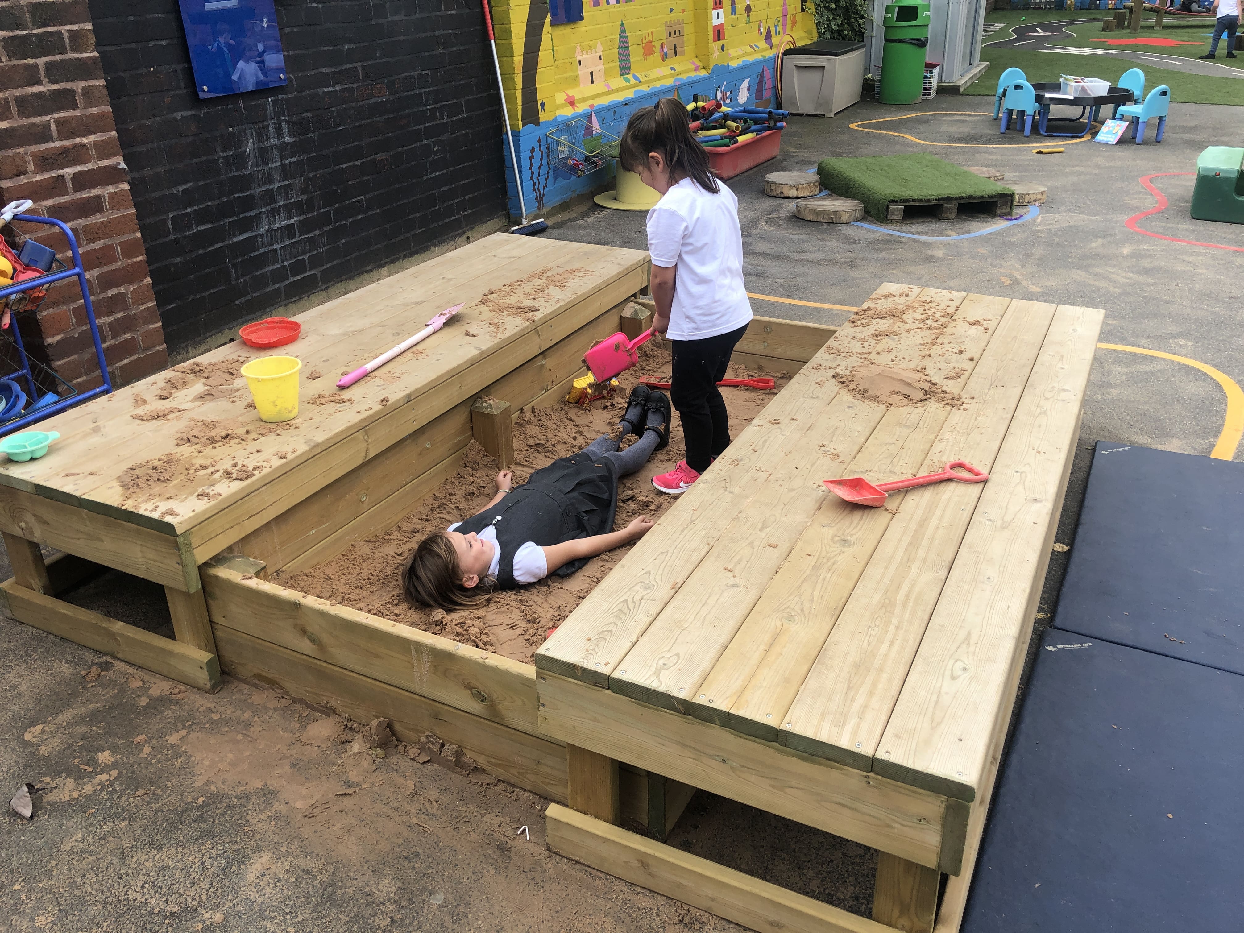 A little girl is lying in the large sand pit, whilst another girl is digging up sand and placing it over the lying girl. They are both smiling and having fun.