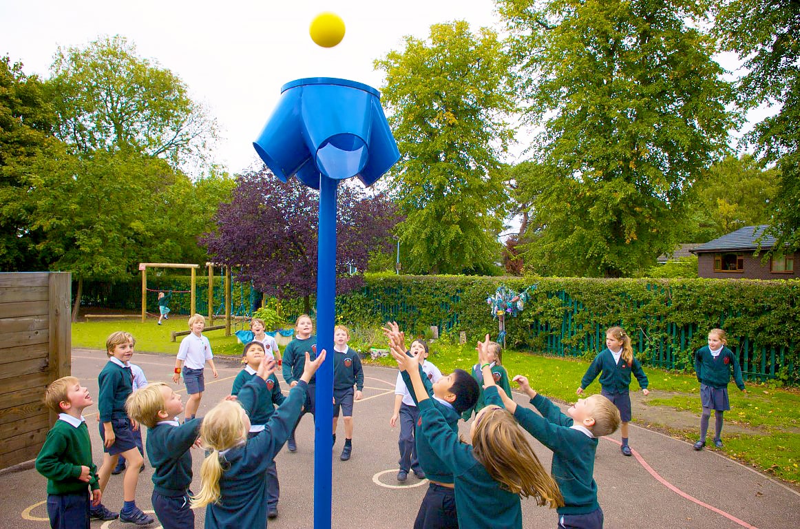 A tall blue chute with a bucket on top is placed in the middle of a playground, which has children throwing balls towards it.