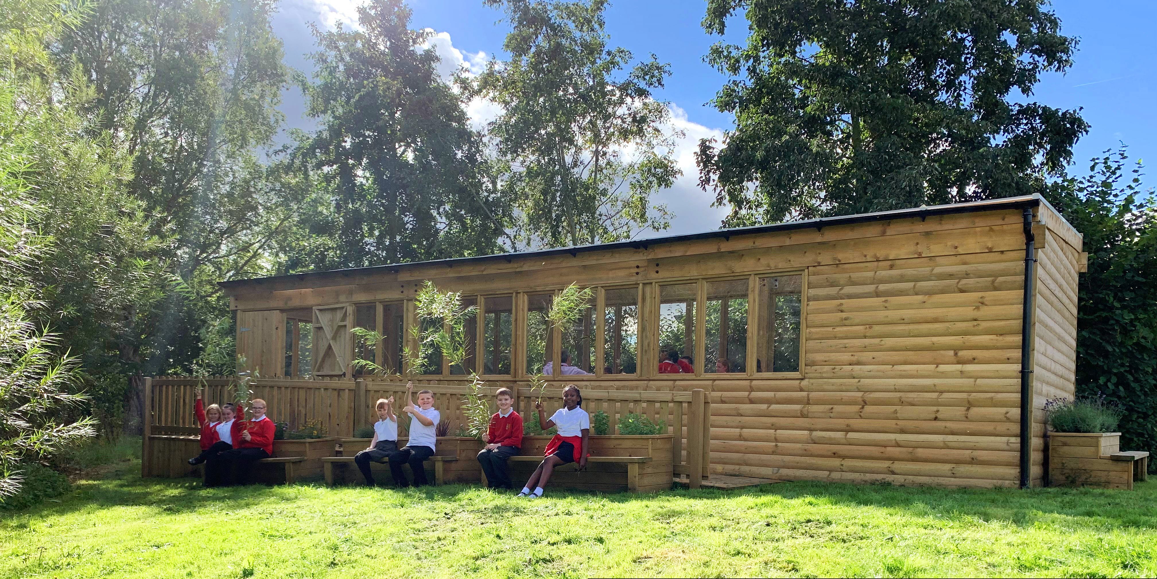 A group of children are sat on benches outside of a wooden playground lodge. The lodge and benches have been placed on top of grass.