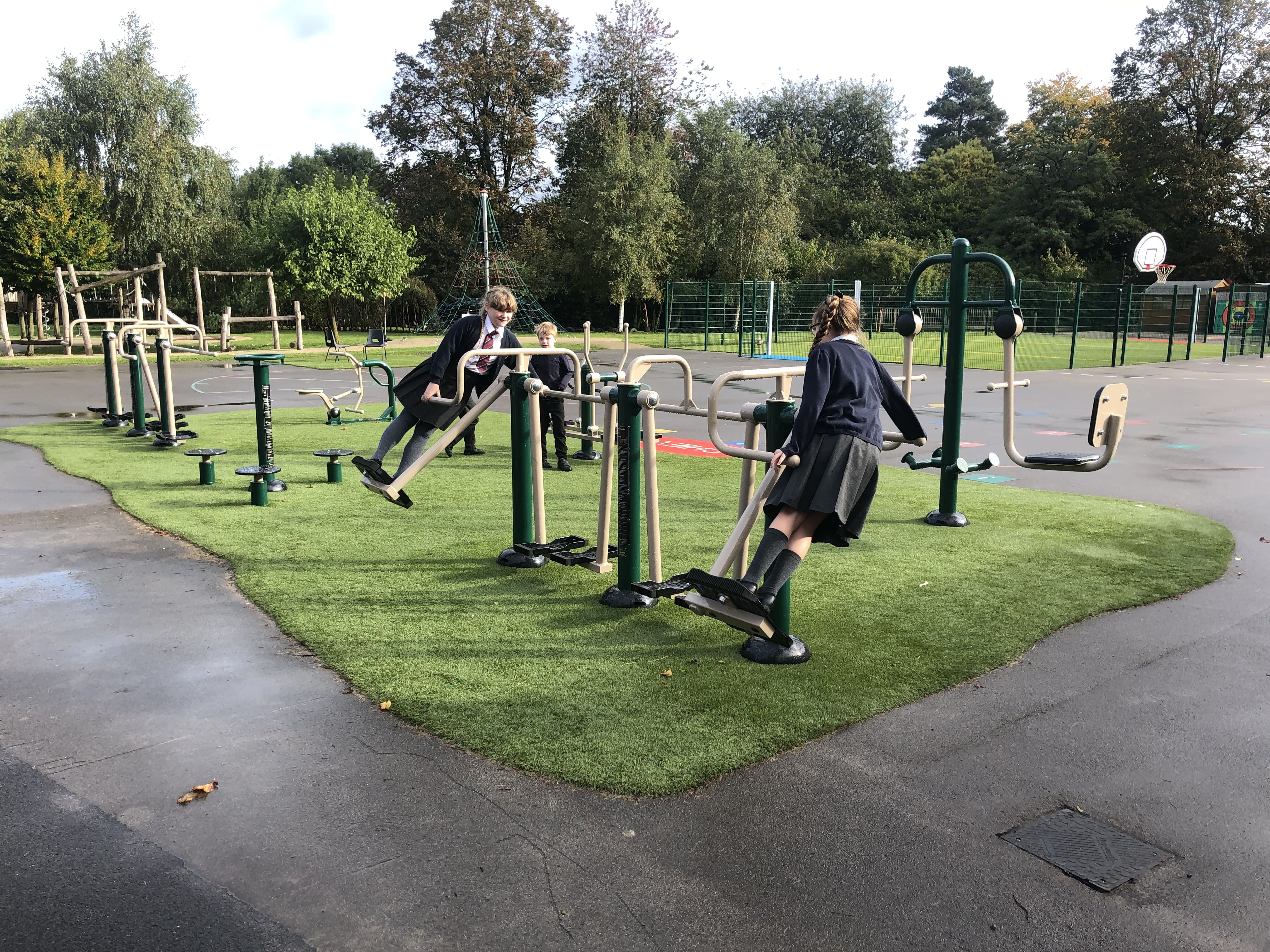 Two girls are rocking on a piece of school outdoor gym equipment, which has been installed on a patch of artificial grass.