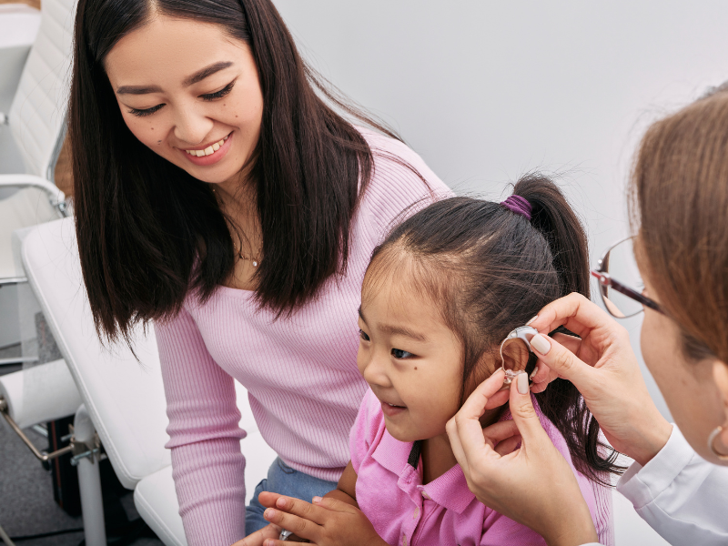 A mother is sat with her daughter as a doctor places a hearing aid on the side of the daughter's head.