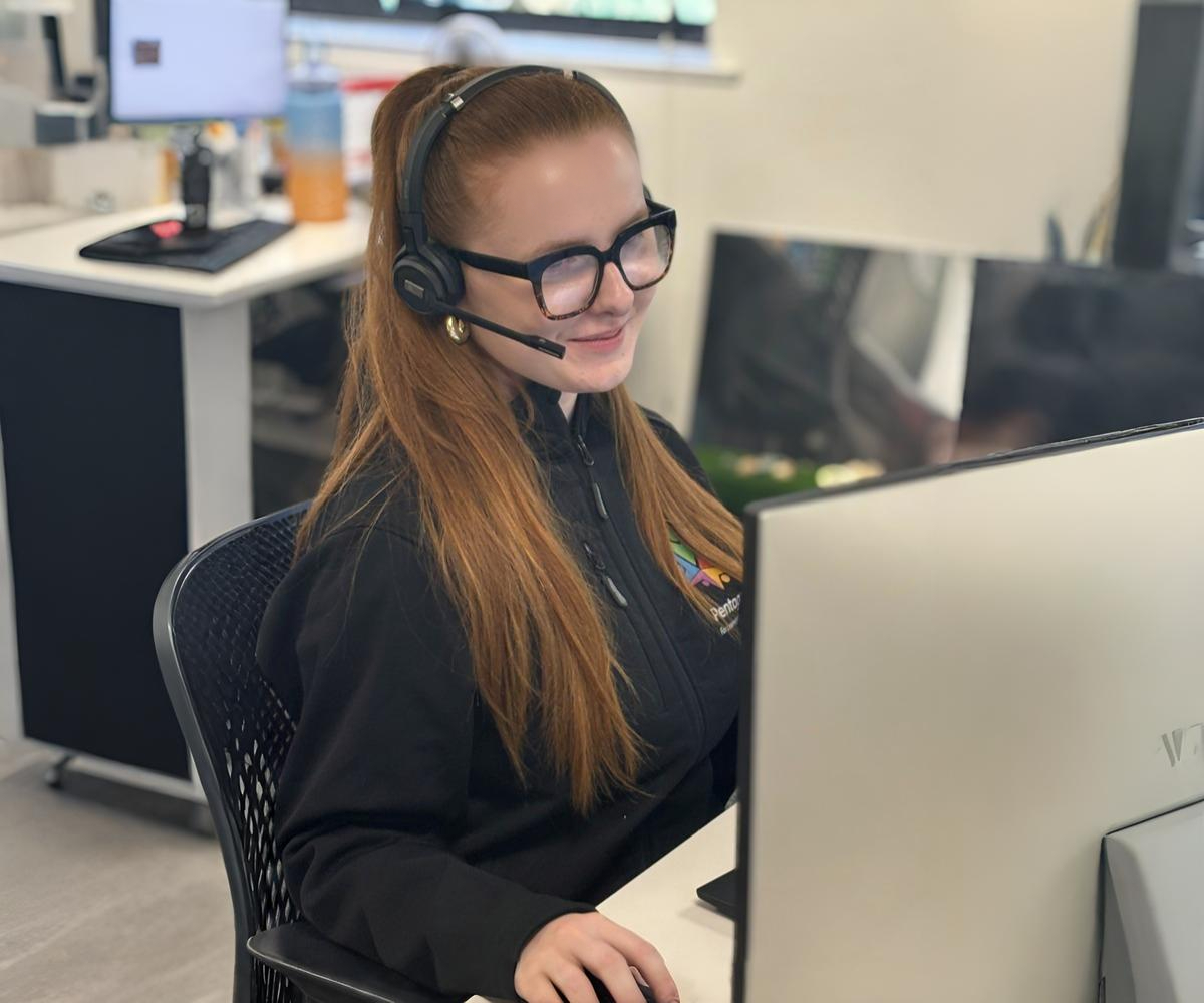 Maddy McGuire is sat at her desk, with a headset on and smiling whilst looking at her computer.
