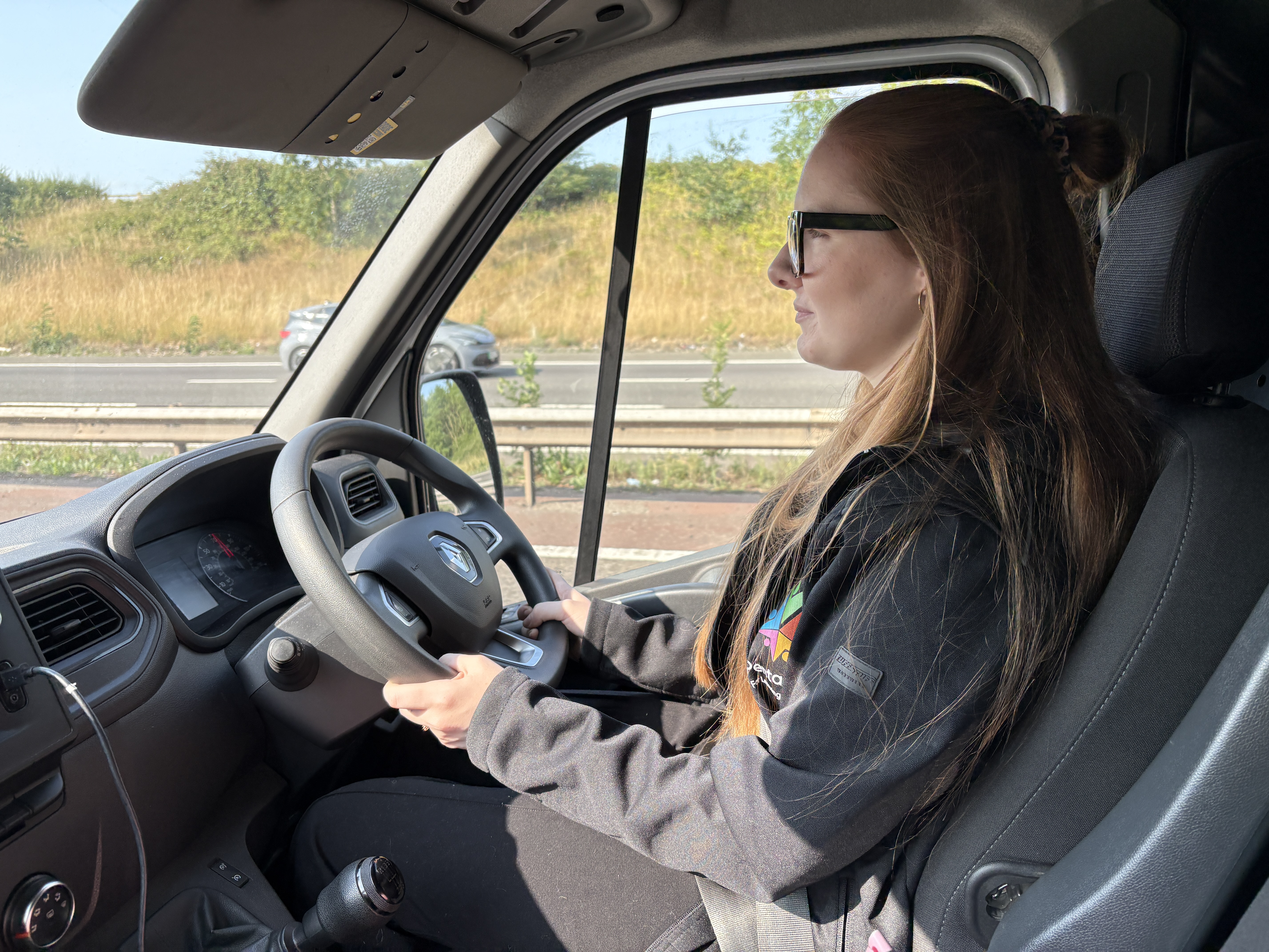 Maddy McGuire is sat inside a van, behind the wheel, driving down a road.