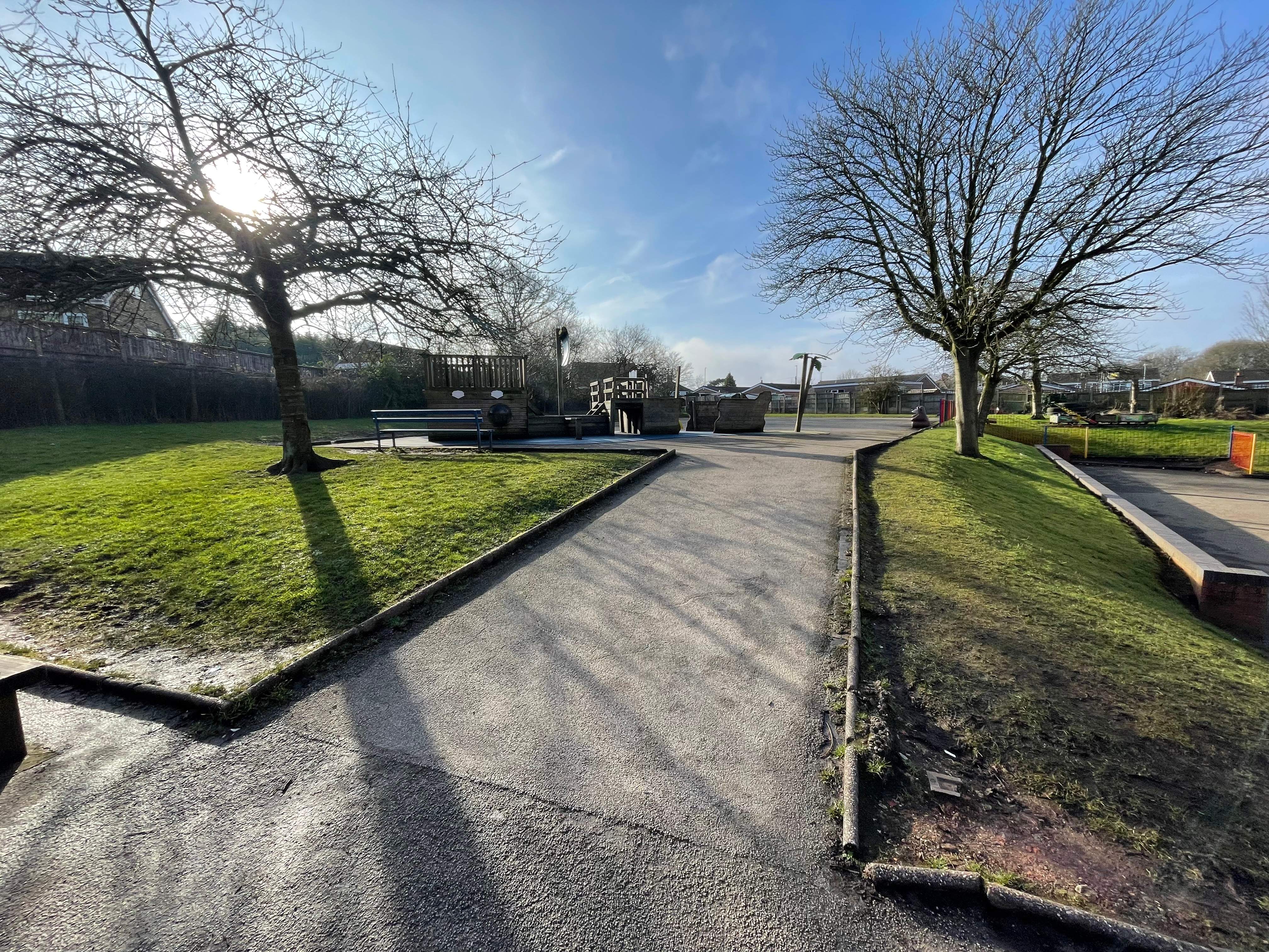 A concrete pathway that leads towards a seating area, with grass on either side with trees.