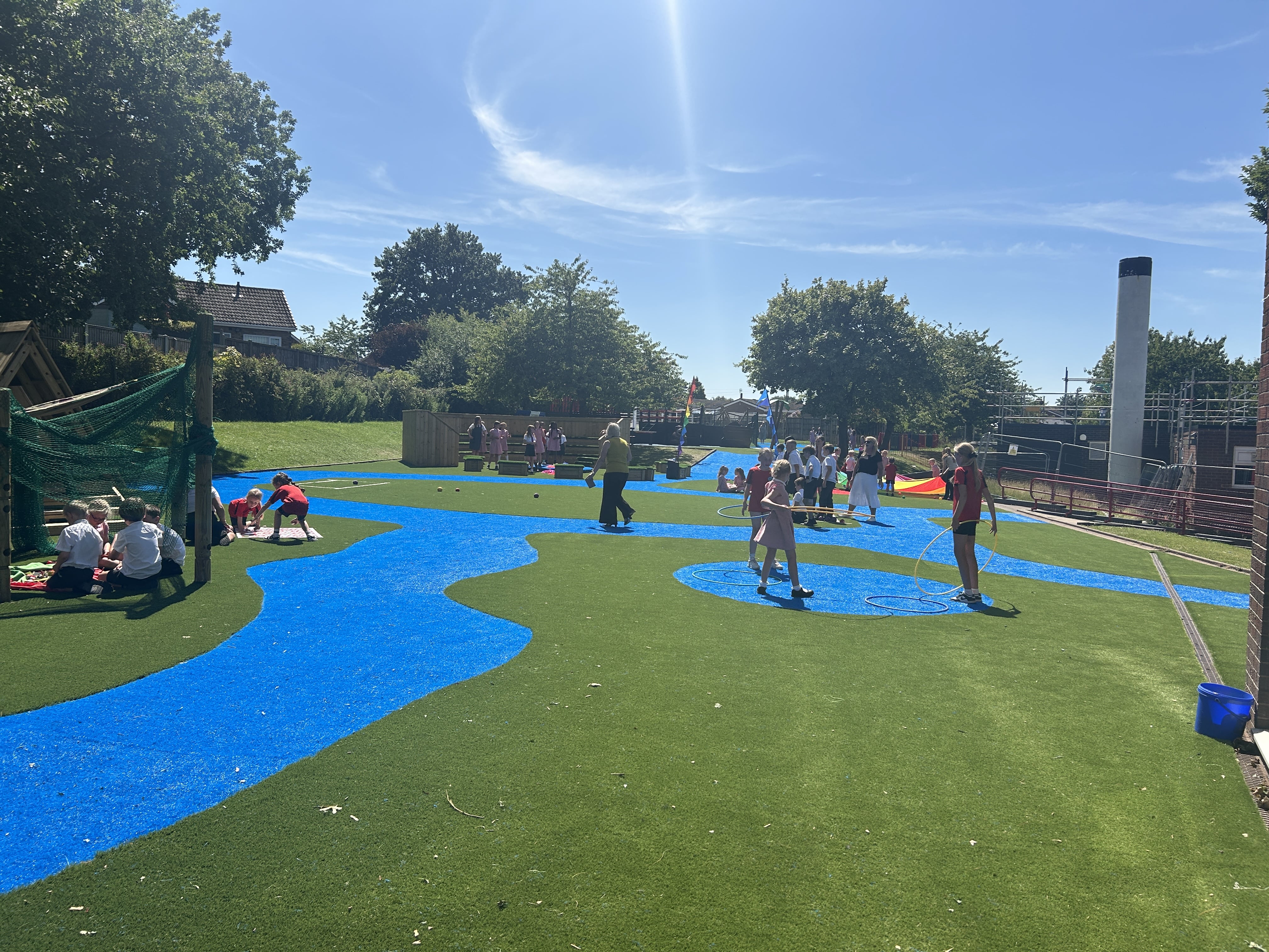 A large group of children are playing with different pieces of play equipment in different areas of the playground as the teacher stands in the centre.