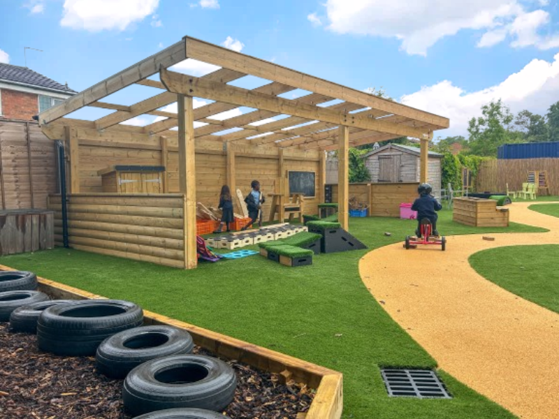 A child is cycling on a wetpour road past an outdoor classroom, which has a few children within it playing with equipment.