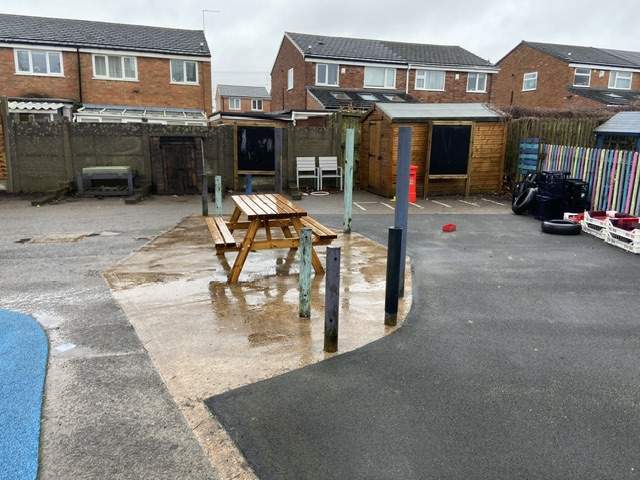 A wet picnic bench on tarmac that is surrounded by worn-out wetpour and fence.