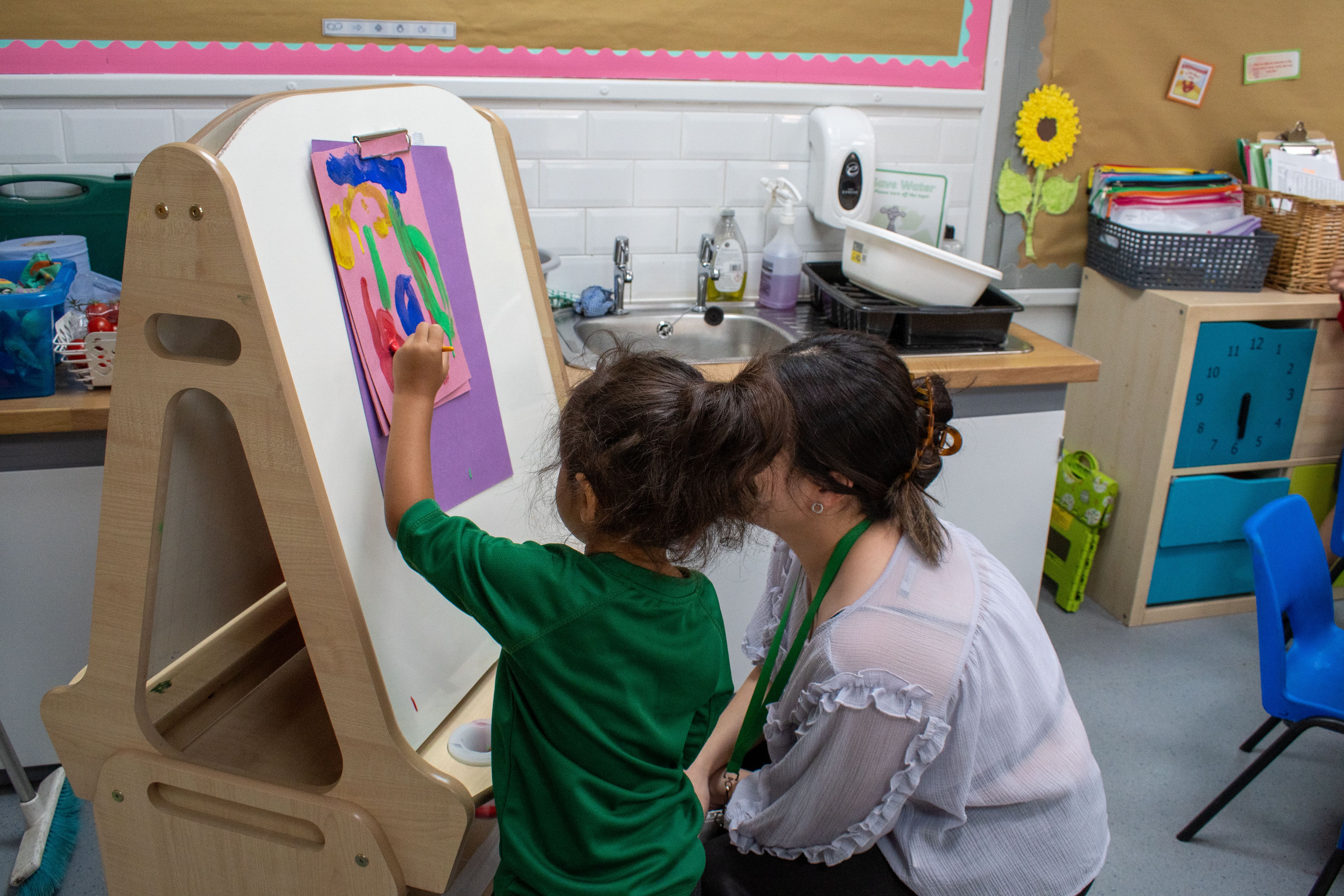 A child and a teacher are painting on a piece of coloured paper that is stuck to a whiteboard easel.