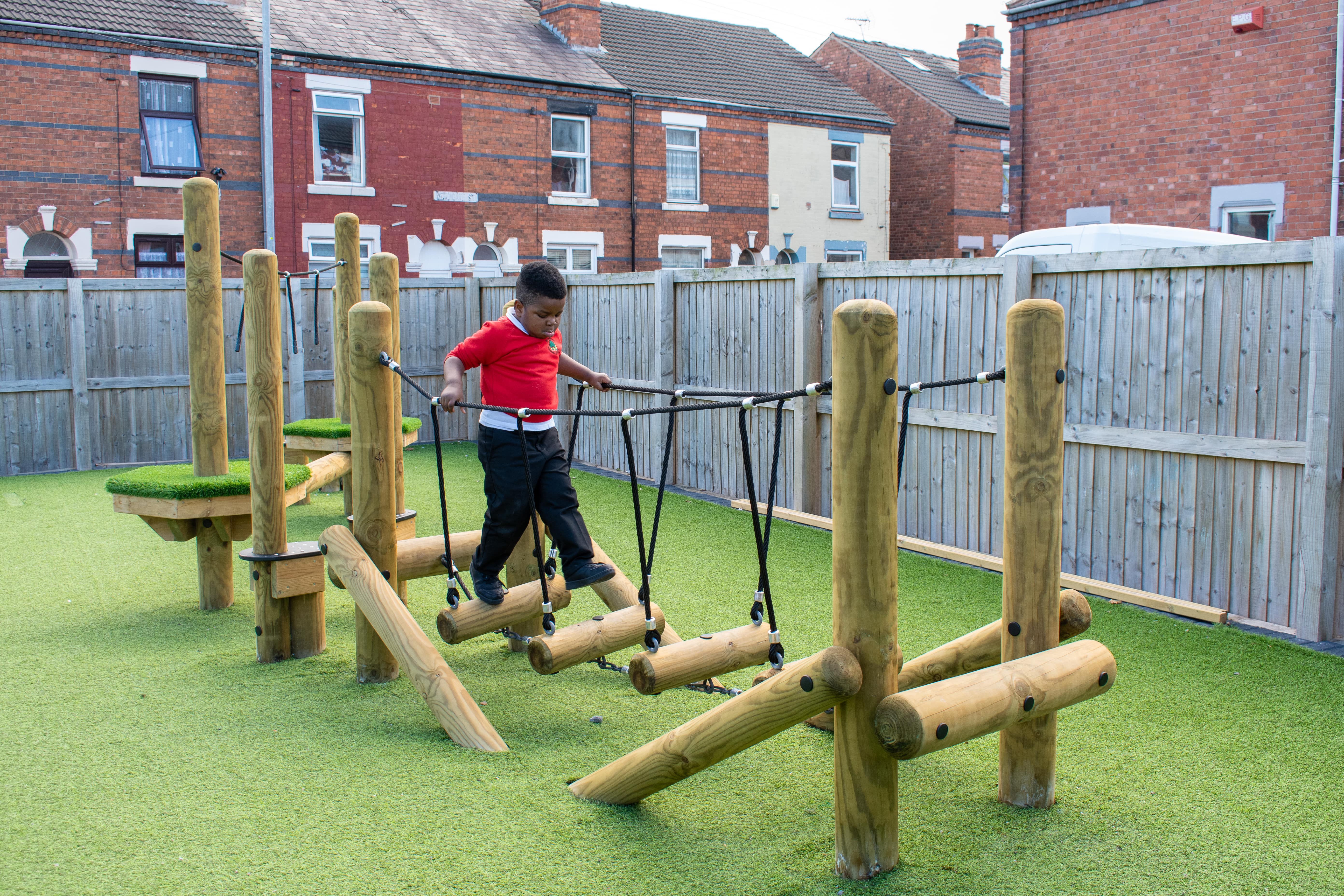 A little boy is crossing a wooden log bridge that has been installed outdoors.