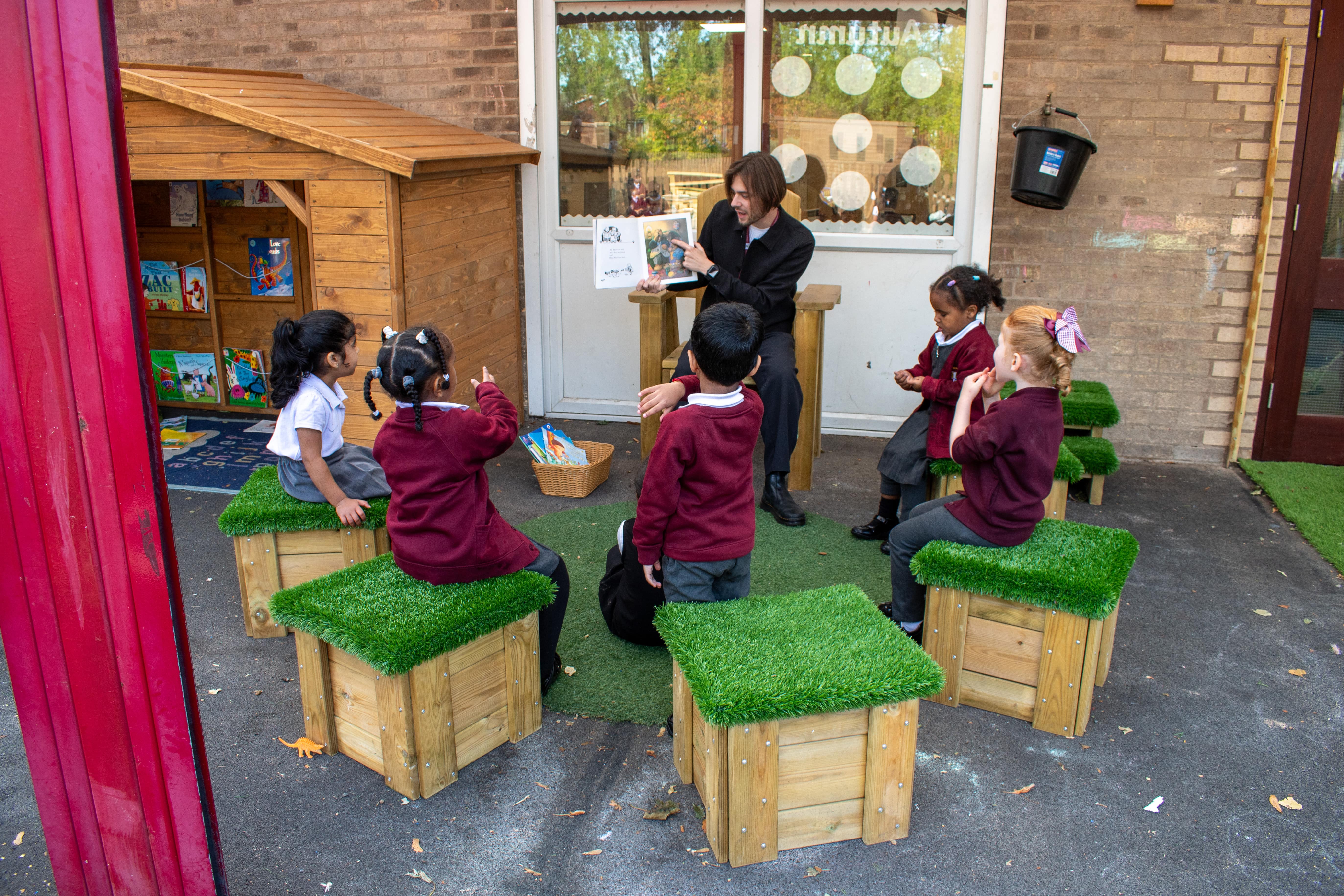 A small group of children are sat on small artificial grass topped blocks that are surrounding a freestanding storytelling chair.