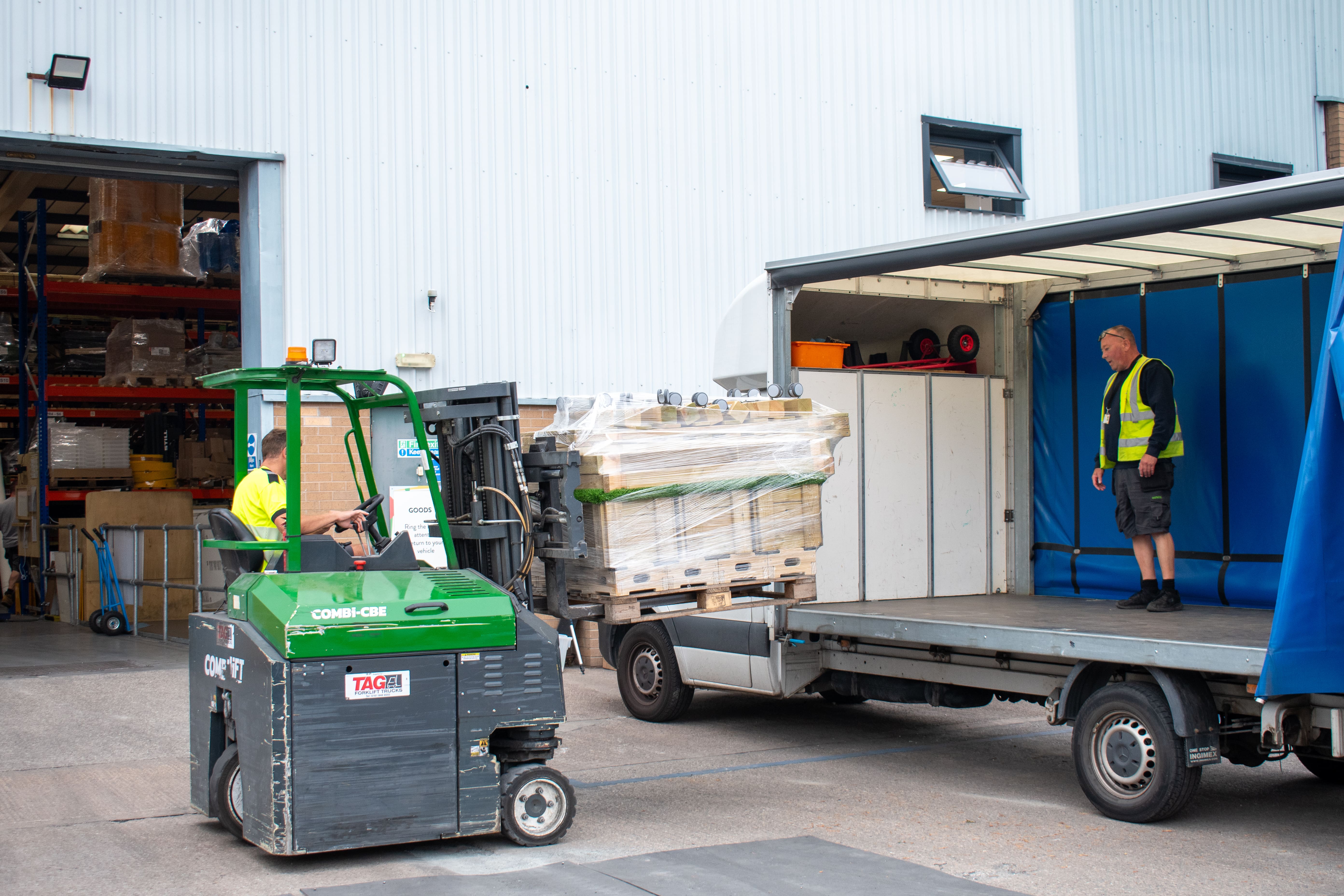 A forklift is carrying a pallet of play equipment over to a delivery lorry, as a man waits to receive it.