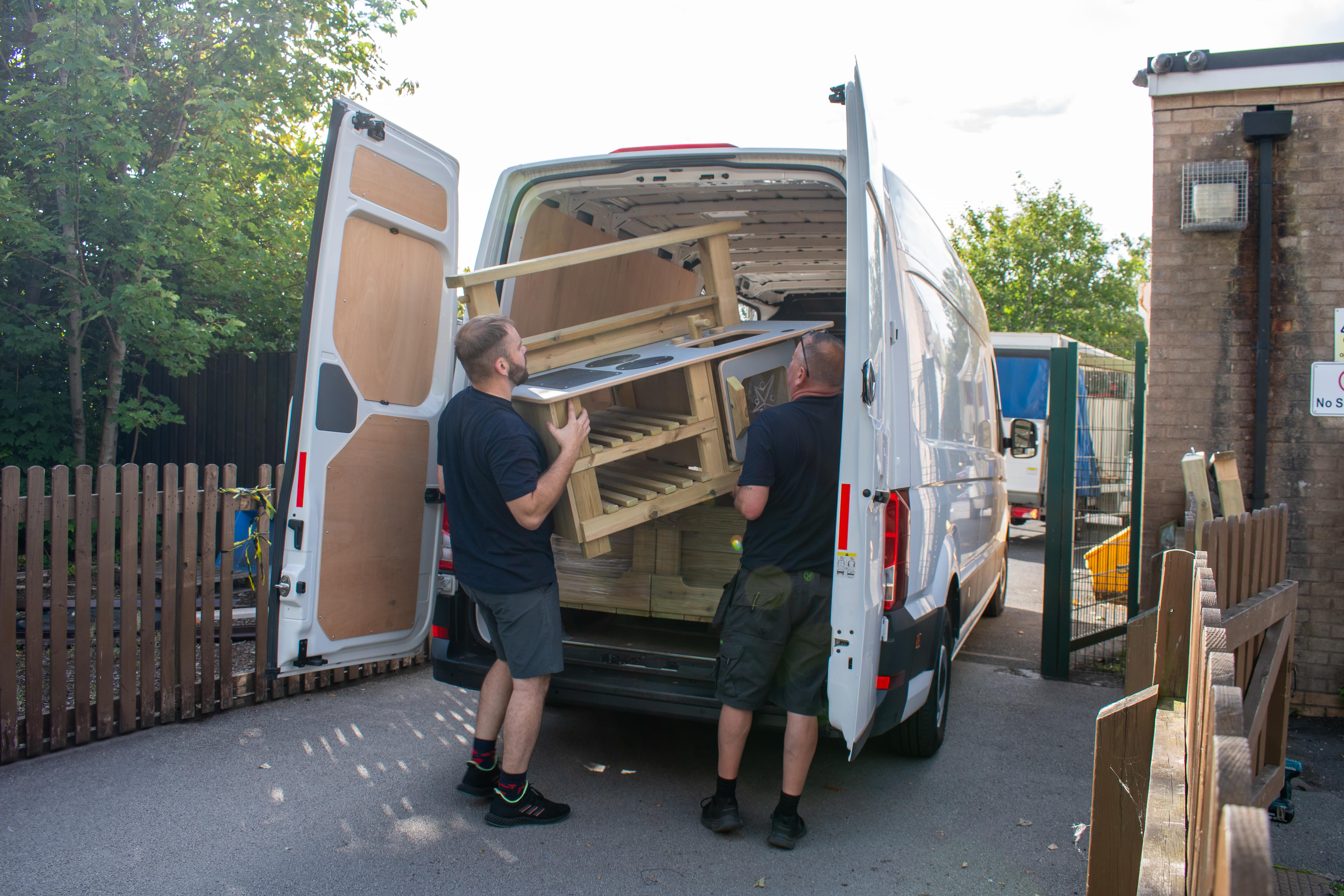 Two men are moving a mud kitchen from the back of a Pentagon Play van to a playground.