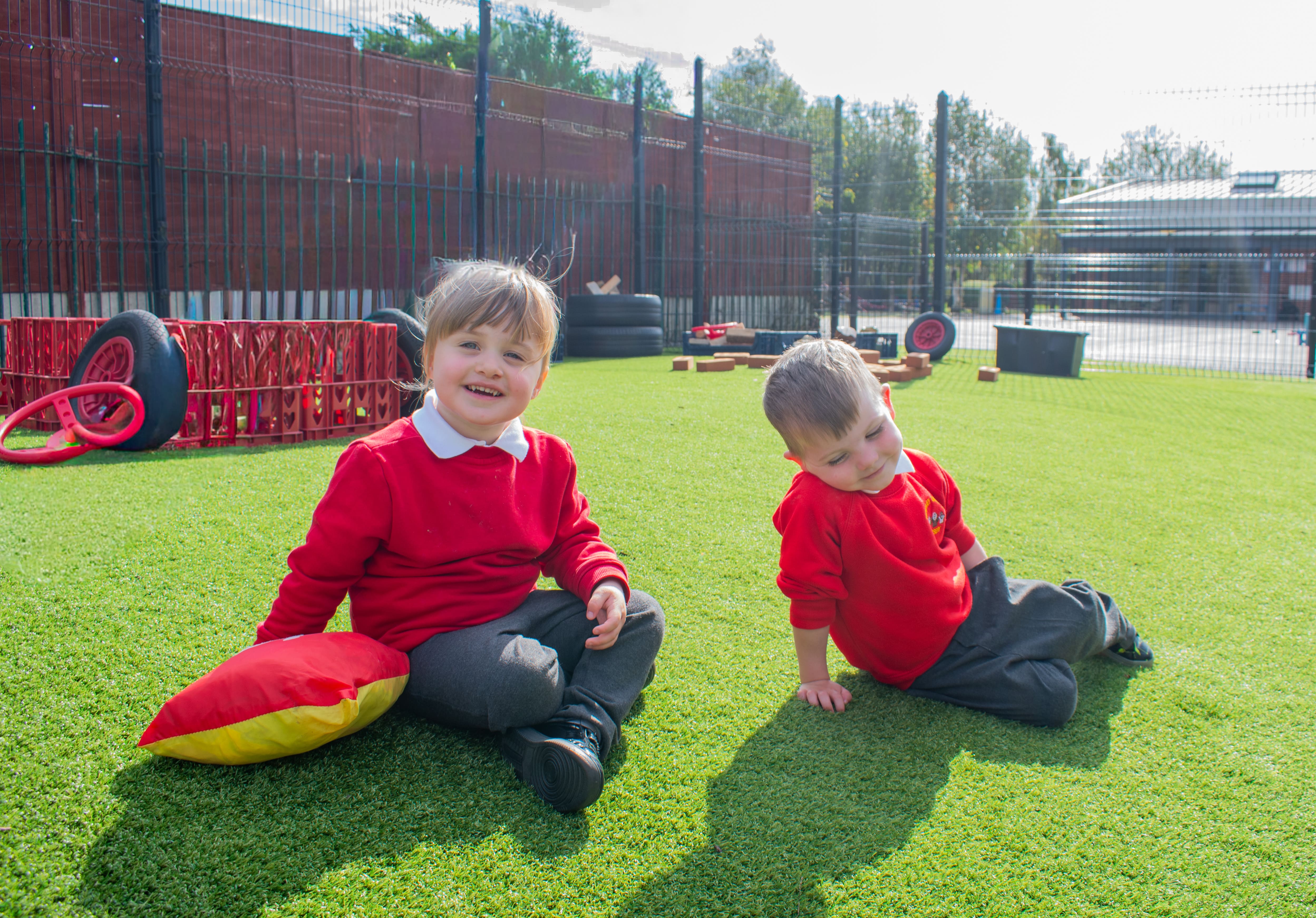 Two children are sat on the artificial grass as they look at the camera and smile.