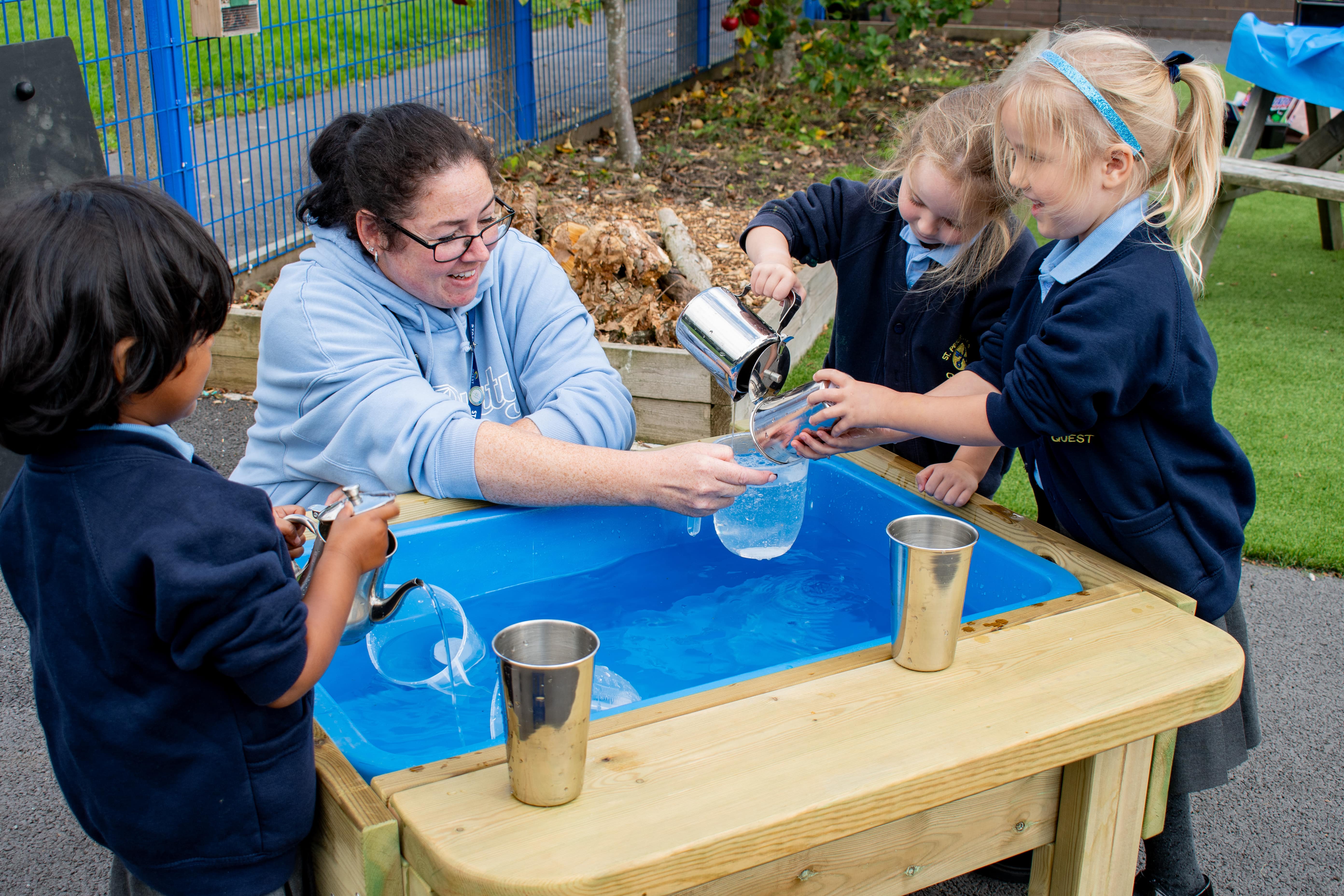 A small group of children are sat with a woman as they play with a water play table.