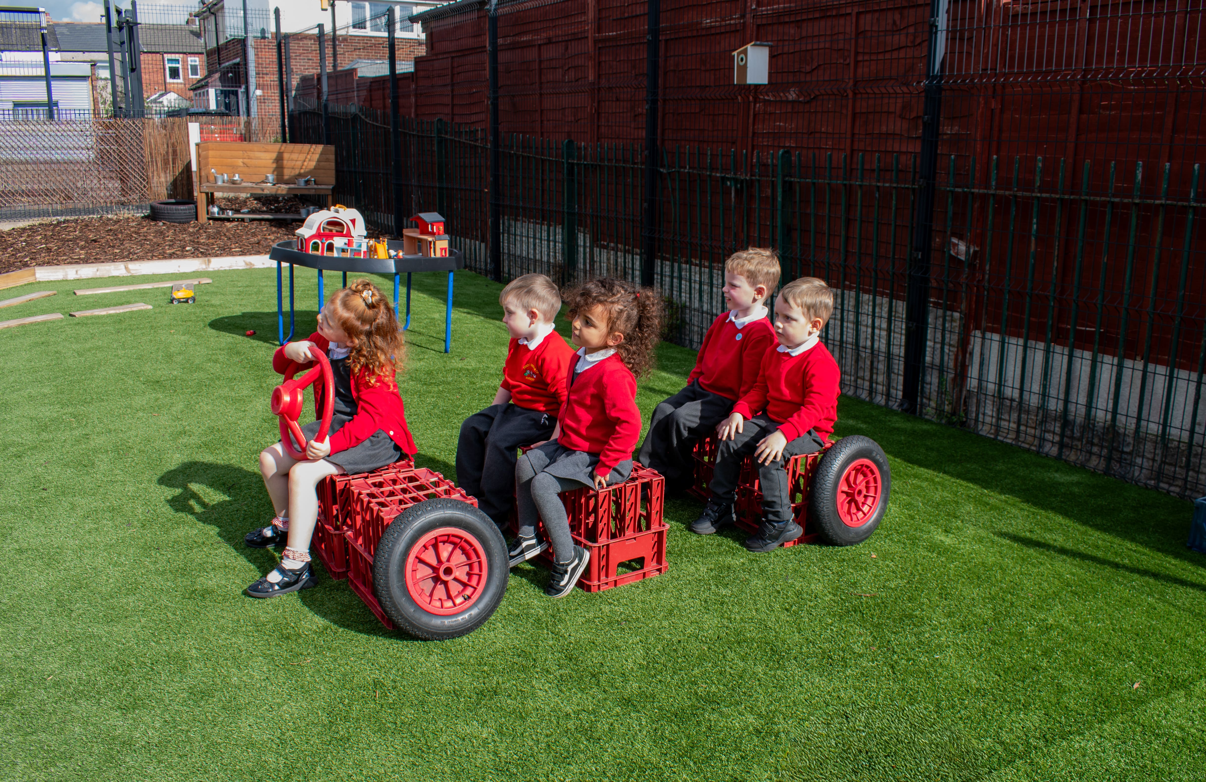 A small group of children are sat on boxes with wheels nearby, creating a fake car that is on top of artificial grass.