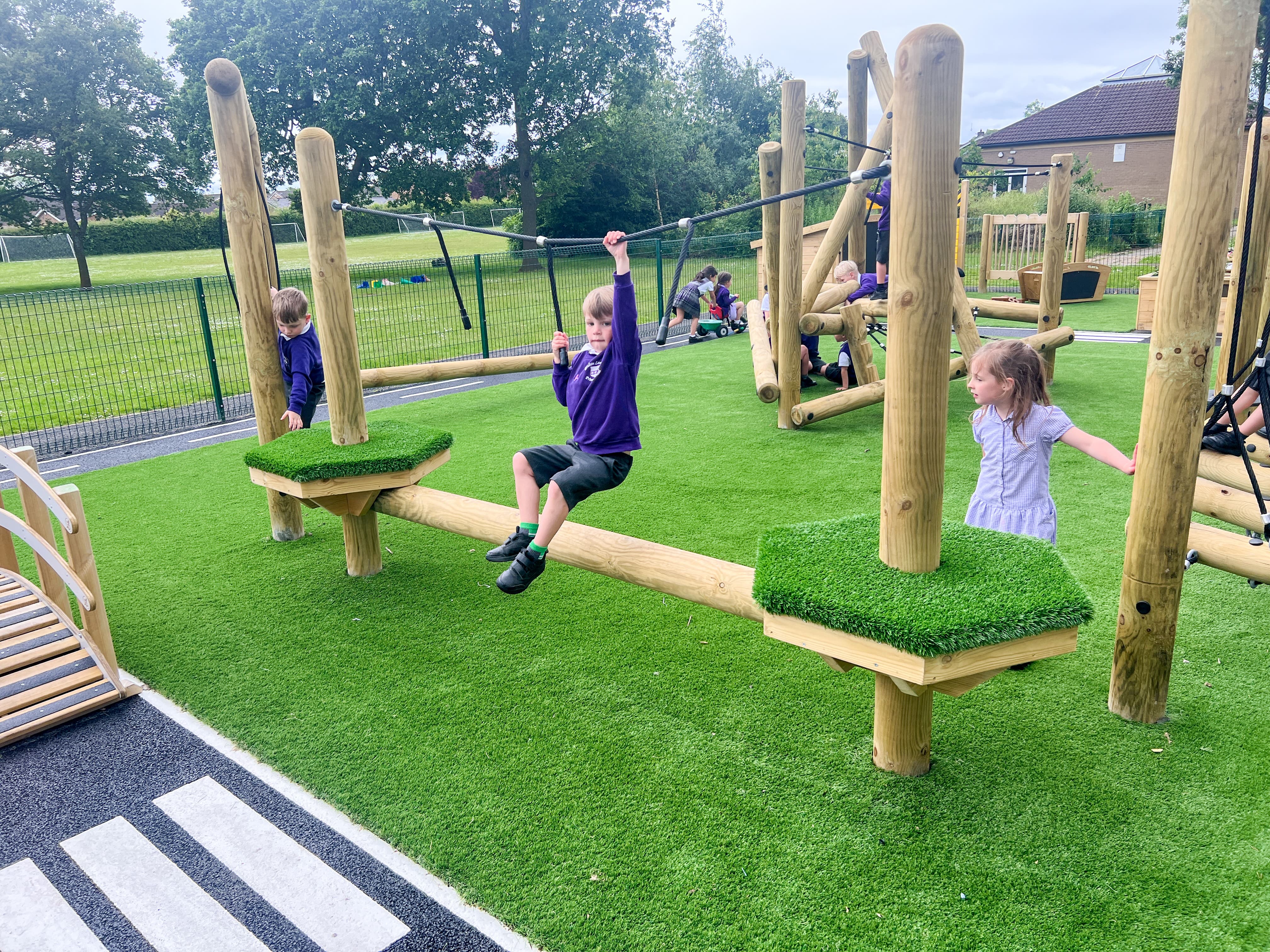 A boy is holding on to a rope as he looks at the camera and smiles, whilst children run around behind, exploring the playground.