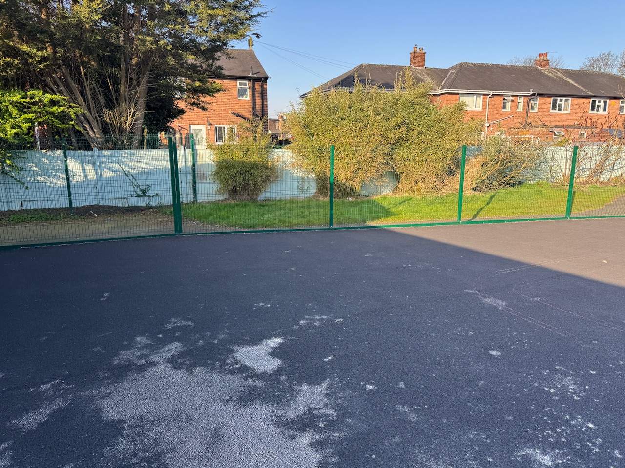 An empty playground that has old tarmac surfacing and green fencing around it.