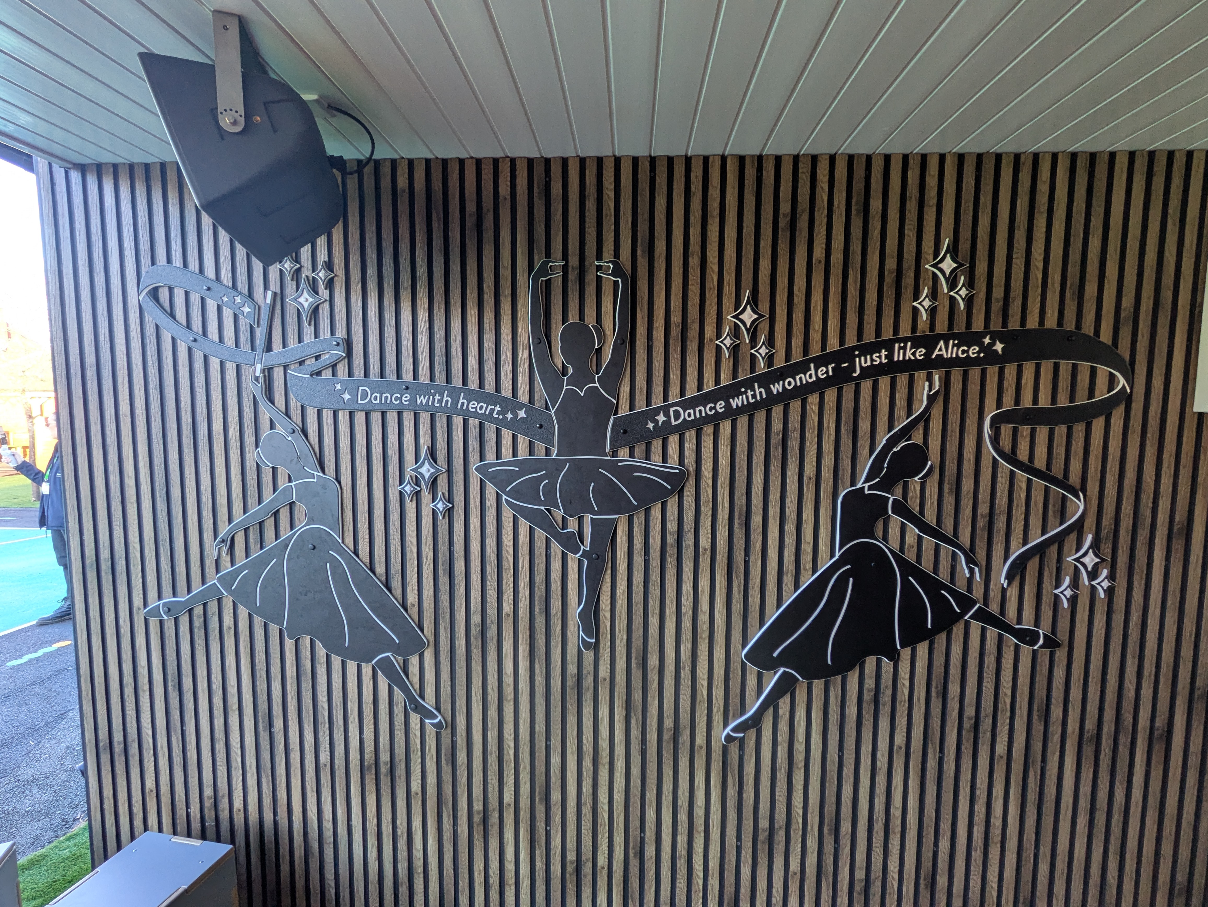 A memorial piece with three ballerinas and a message saying "Dance with Heart. Dance with wonder - just like Alice" in a ribbon.