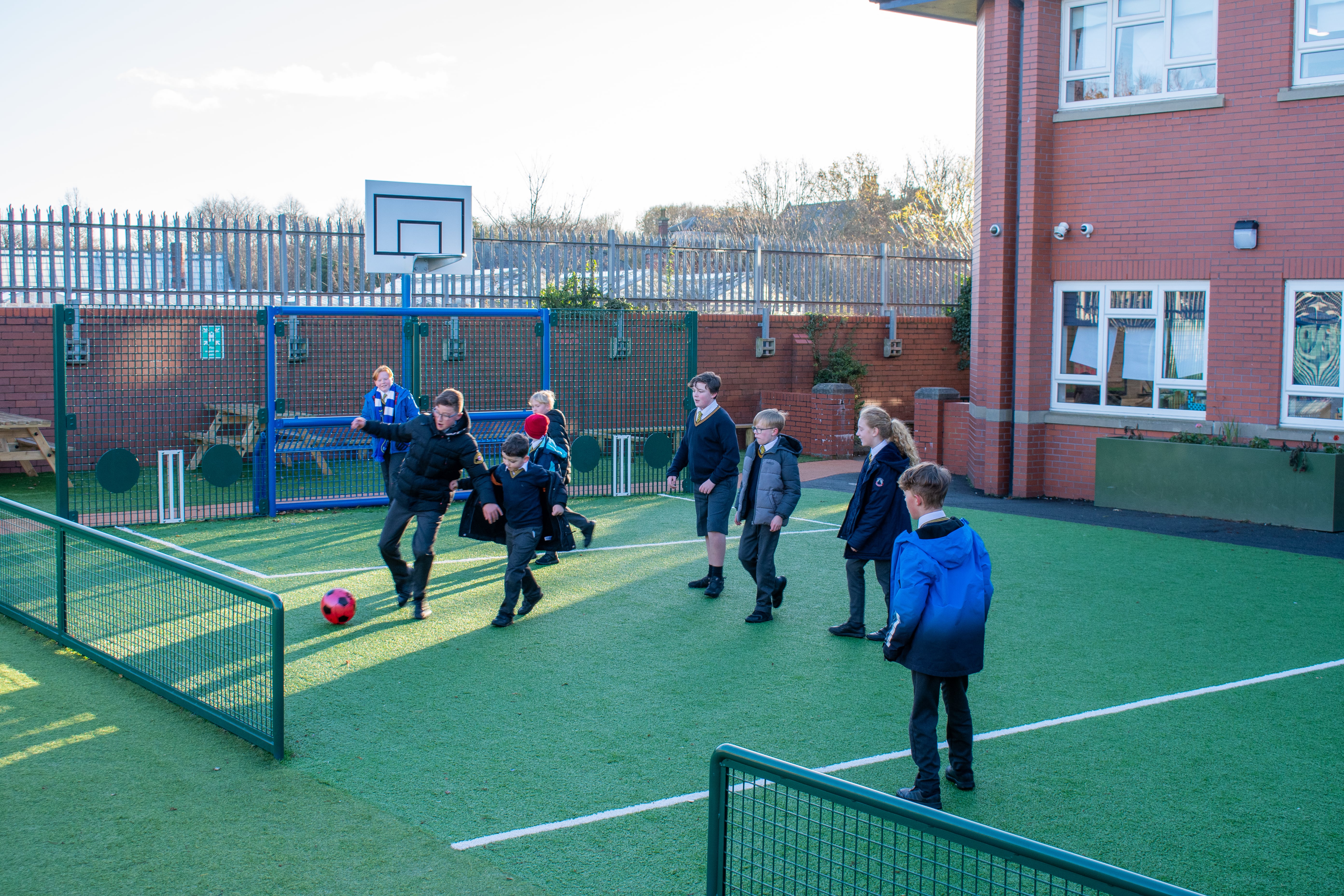 A MUGA pitch with a group of children playing football on it.